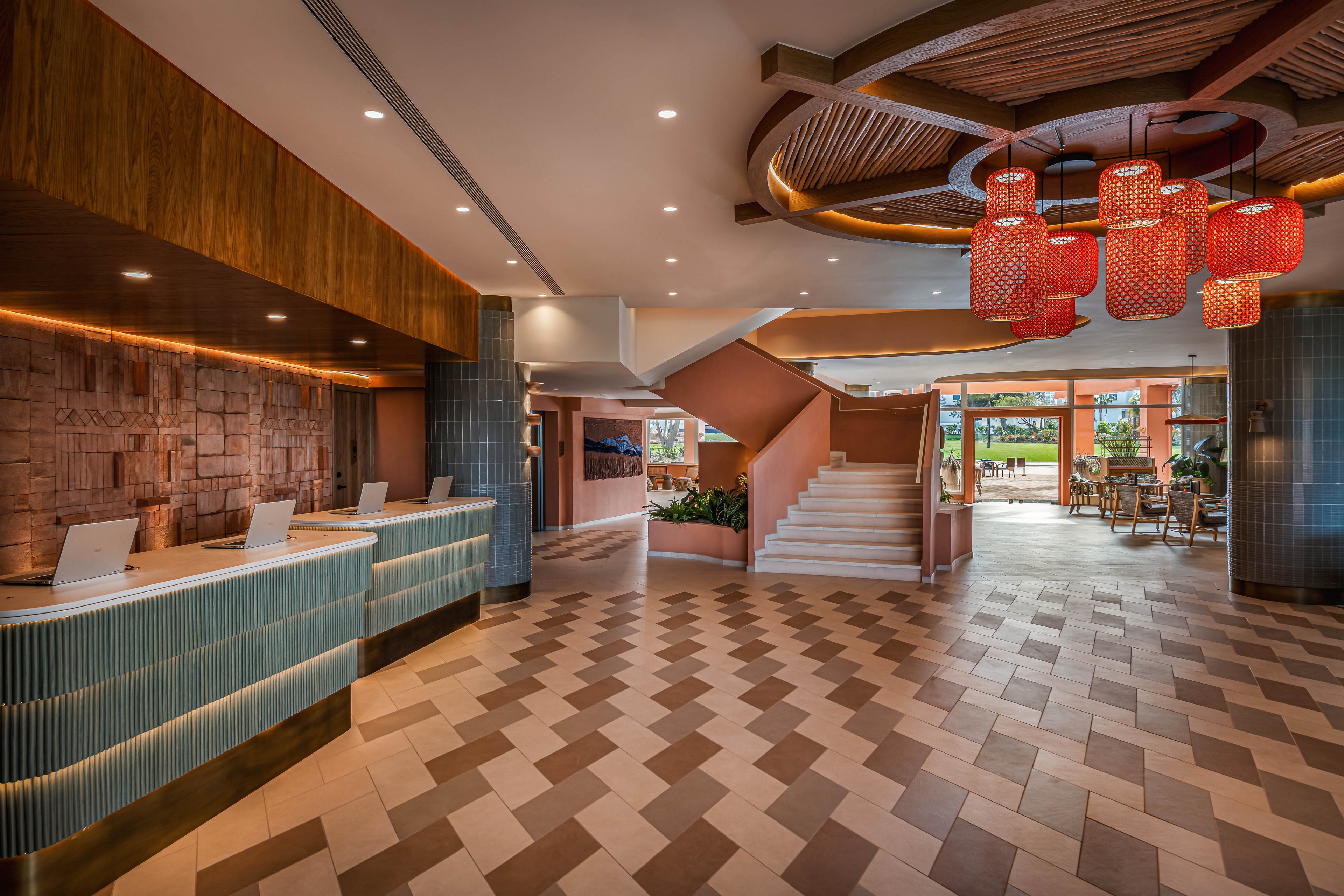 Spacious contemporary hotel lobby featuring a long reception desk with multiple check-in stations. The setting includes a patterned tile floor leading toward a central staircase and open seating area. Warm wooden accents, teal tiles, and large red hanging lanterns create a welcoming atmosphere. Natural light enters from large windows at the far end, highlighting the clean, modern interior design.