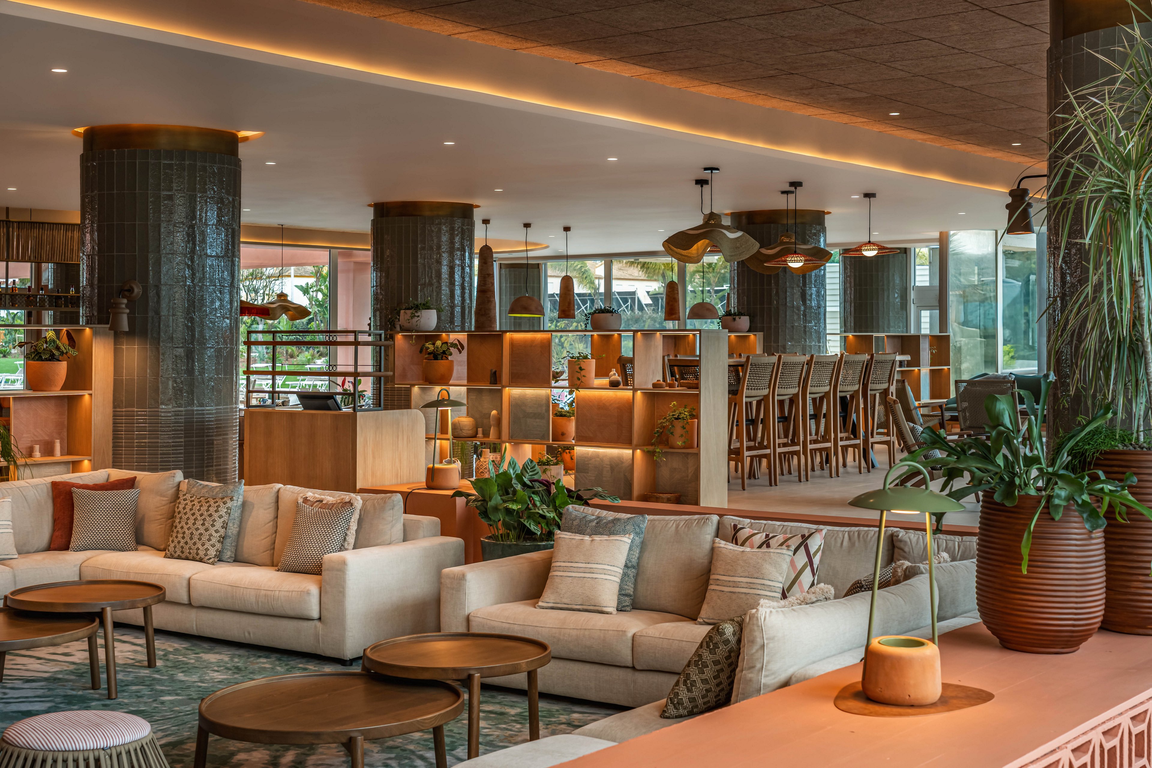 A spacious modern lounge features cream-colored sofas and wooden coffee tables arranged in a cozy seating area. The setting appears to be a hotel or resort lobby with a bar and high stools visible in the background. Warm ambient lighting highlights textured columns, open shelving, and decorative plants throughout the space. Earthy tones of beige, terracotta, and green create a relaxed, inviting atmosphere suitable for socializing or unwinding.