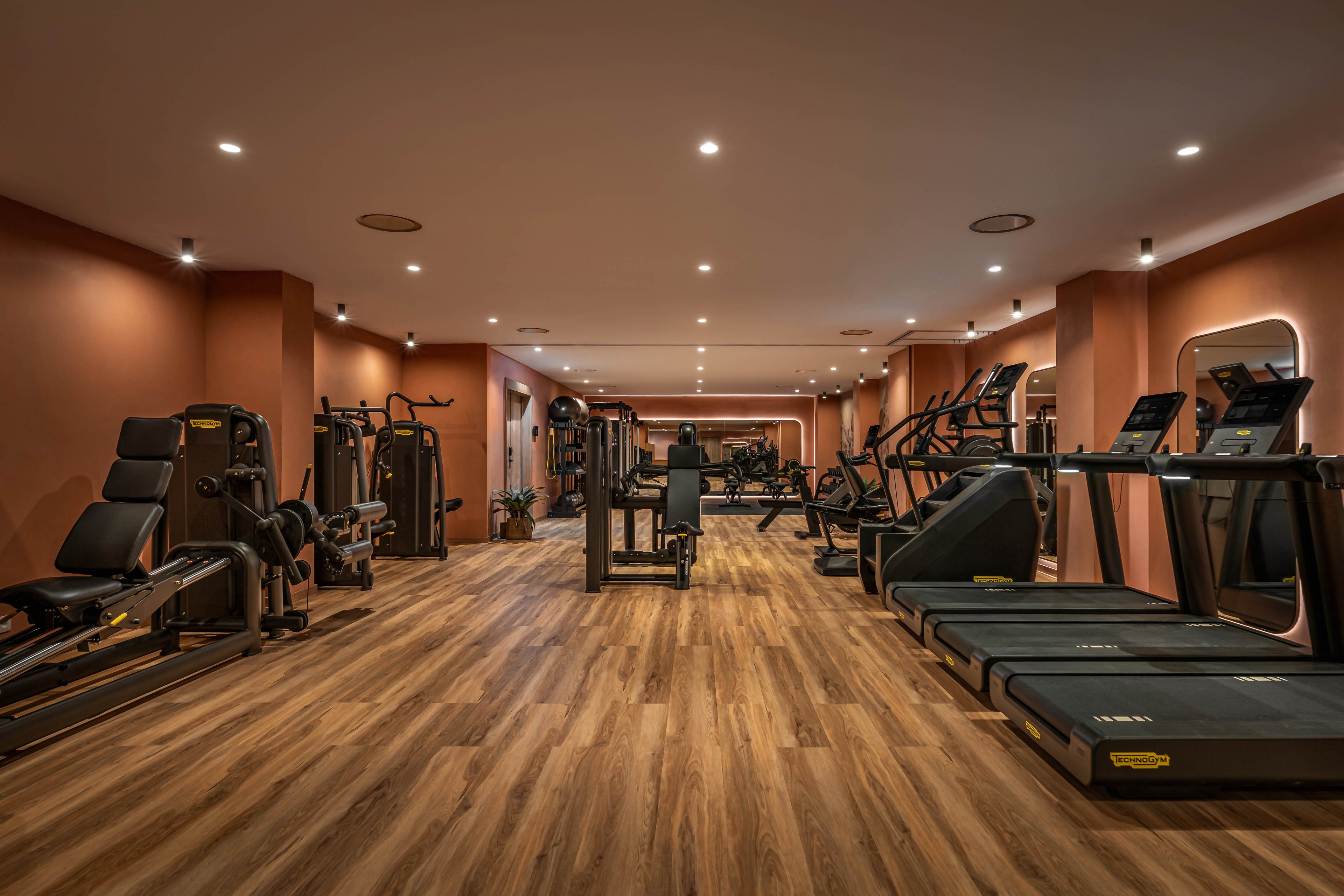 A spacious modern gym interior features rows of treadmills, ellipticals, and strength machines neatly arranged on a wooden floor. Warm recessed lighting in the ceiling creates an inviting atmosphere against the muted terracotta walls. The room is empty of people, emphasizing the clean, organized layout and high-end fitness equipment. Large mirrors and open floor space enhance the sense of depth and comfort in the workout area.
