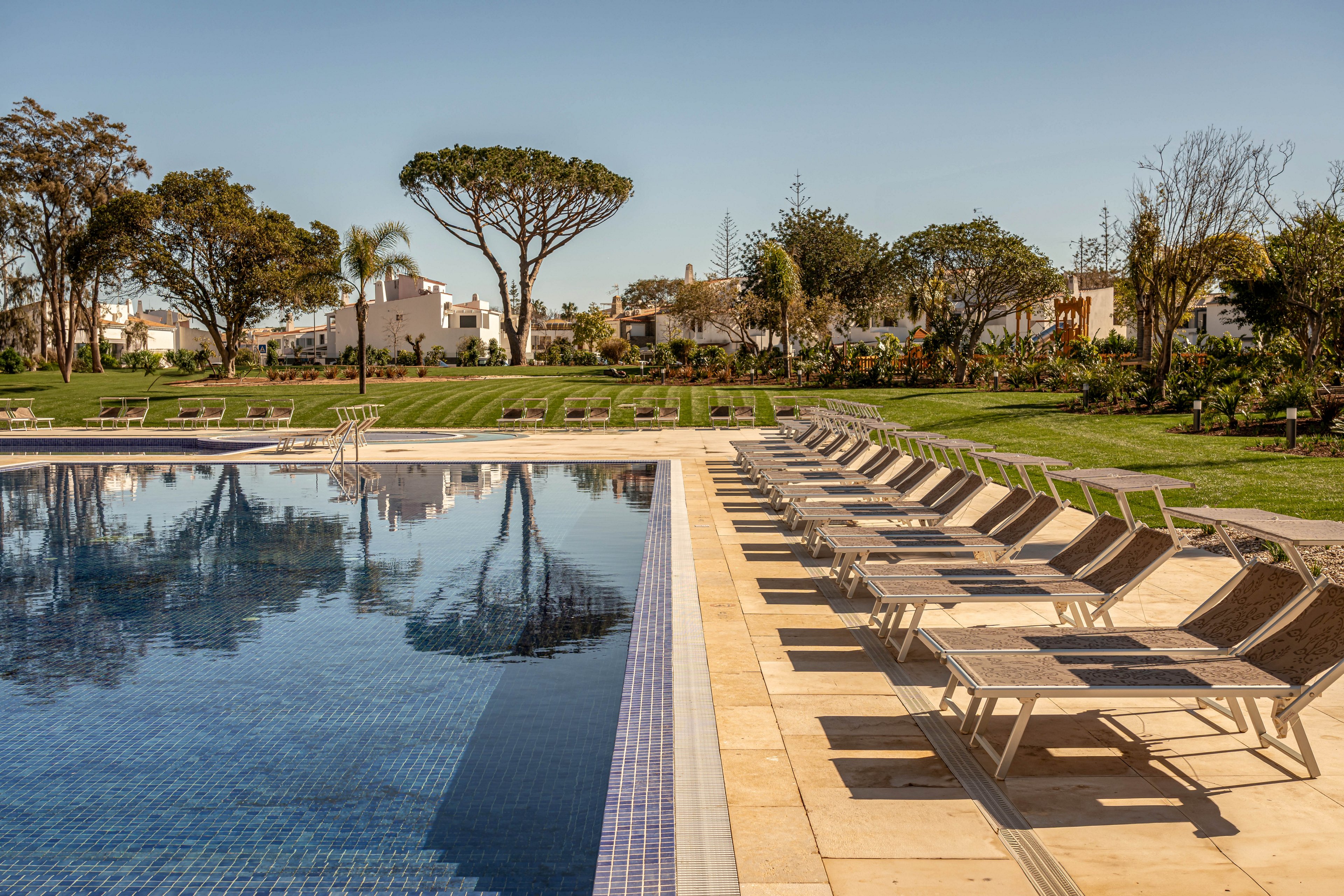 A large outdoor swimming pool sits beside a long row of empty sun loungers. The scene is set in a landscaped resort garden with neatly trimmed grass and various trees and shrubs. Reflections of the sky and surrounding greenery appear on the calm pool water. Residential-style buildings are visible in the background under a clear blue sky, suggesting a peaceful vacation setting.
