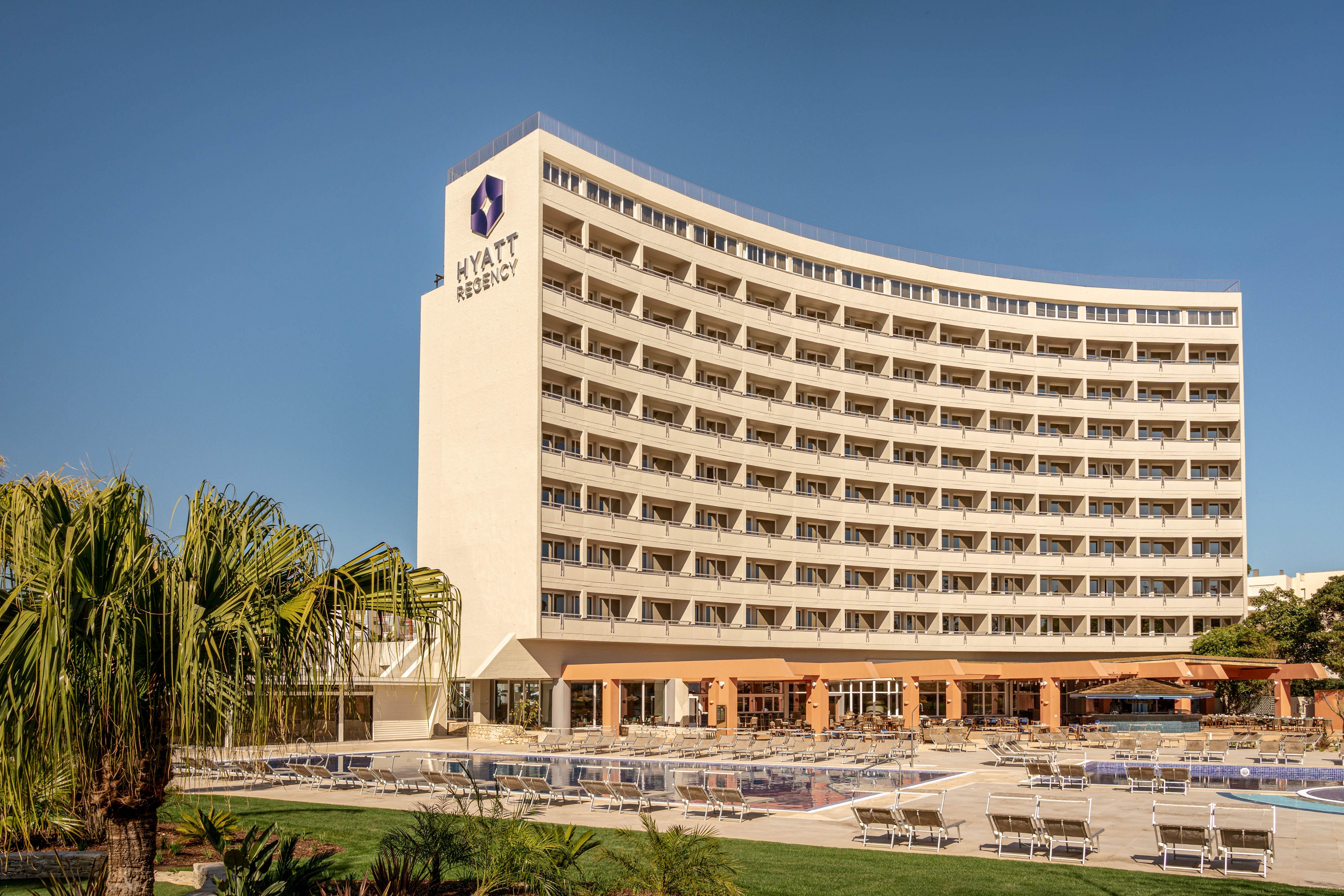 A large curved Hyatt Regency hotel building stands prominently under a clear blue sky. The multi-story structure features rows of balconies facing an outdoor pool and sunbathing area. Numerous lounge chairs and umbrellas are arranged neatly around the pool, suggesting a resort atmosphere. Palm trees and landscaped greenery frame the foreground, emphasizing a warm, coastal vacation setting.