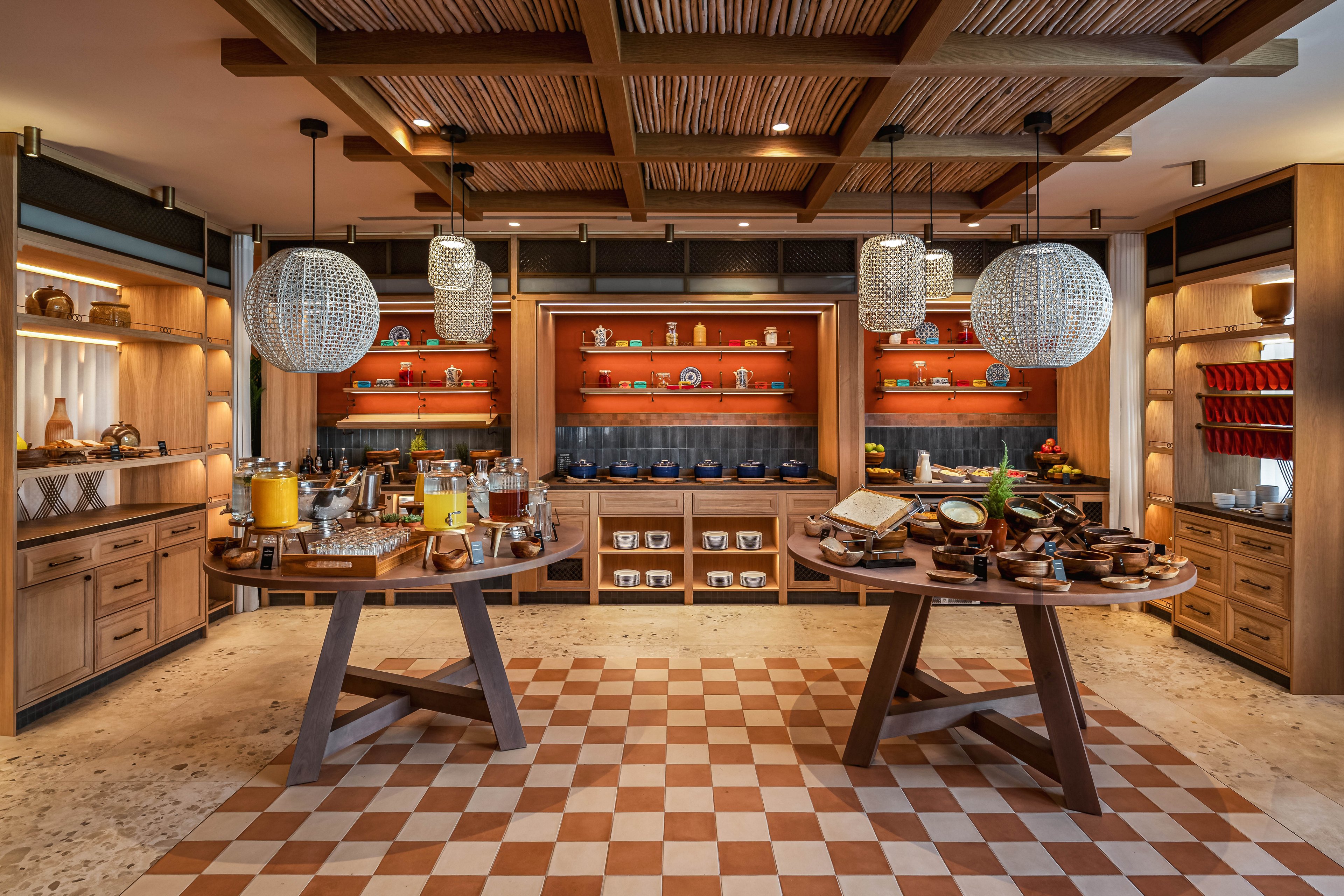 A stylish breakfast buffet is arranged in a warm, wood-paneled dining area. Two round tables display an assortment of cereals, pastries, fruits, and beverages in glass jars and serving dishes. The background features open shelving with neatly arranged plates, bowls, and decorative pottery, highlighted by soft ambient lighting. Large patterned pendant lights hang from a wooden ceiling above a checkered tile floor, creating an inviting and upscale atmosphere.