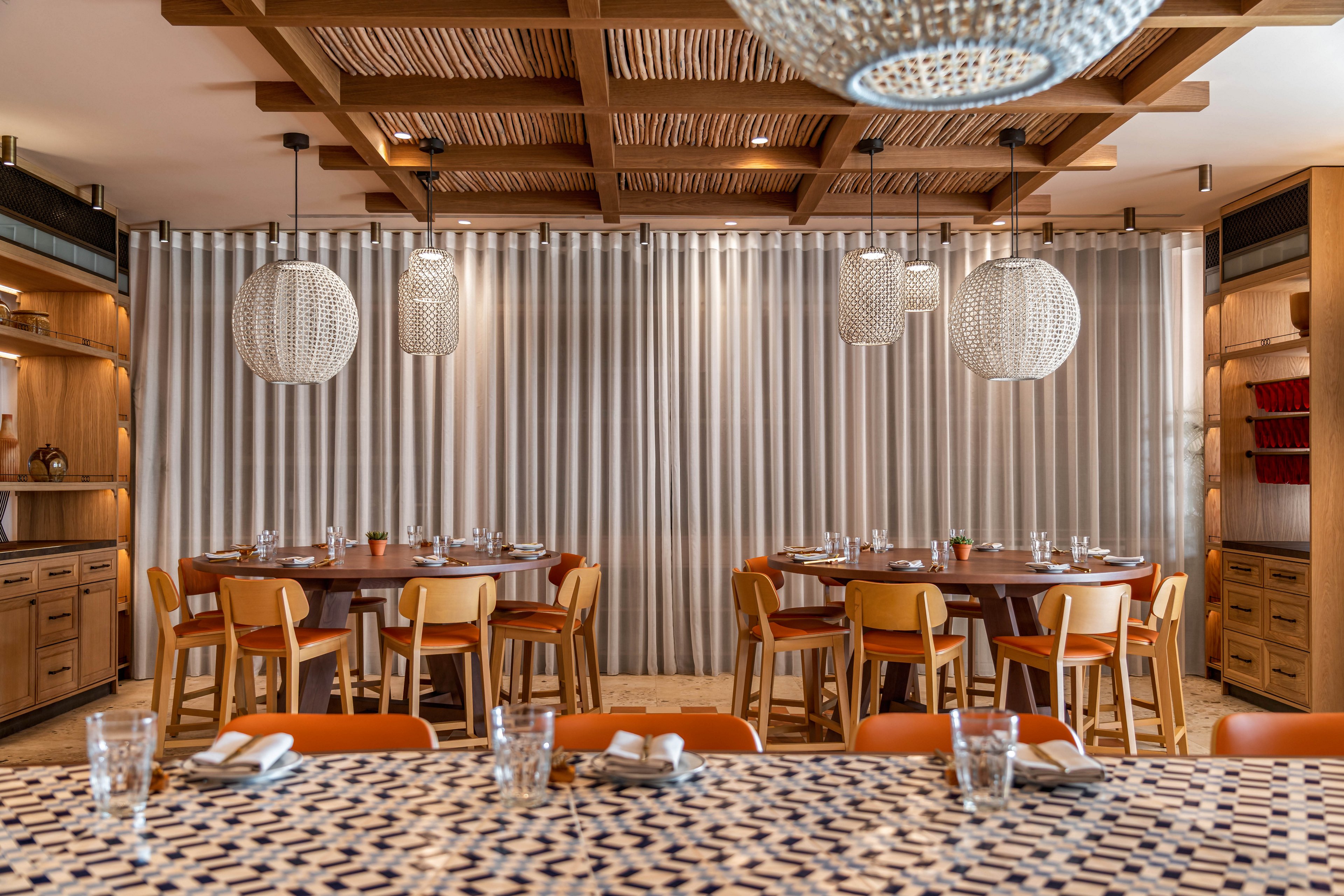A contemporary restaurant dining room features several round wooden tables set with glassware and neatly folded napkins. Warm wooden chairs surround the tables, and built-in shelving with decor accents lines the side walls. Large woven pendant lights hang from a wooden slat ceiling, casting a soft glow over the space. A patterned foreground surface adds visual interest, while neutral curtains create a calm, intimate backdrop.