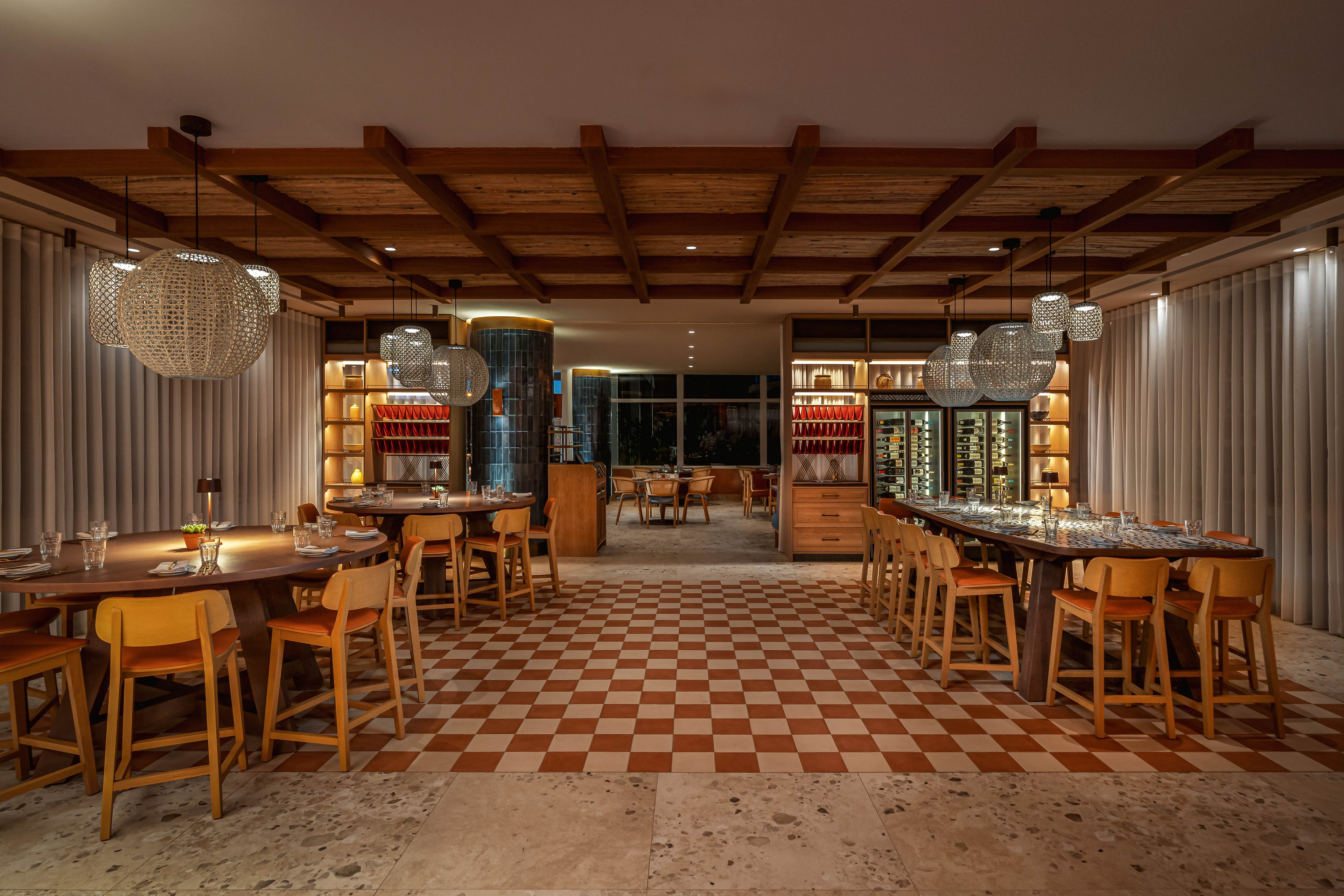 Spacious restaurant interior featuring long wooden tables and neatly arranged chairs. The setting is warmly lit with pendant lights and illuminated shelving displaying bottles and glassware. A checkered tile floor leads toward refrigerated wine cabinets and a central service area at the back. The overall atmosphere feels cozy, refined, and inviting for group dining or wine tasting.