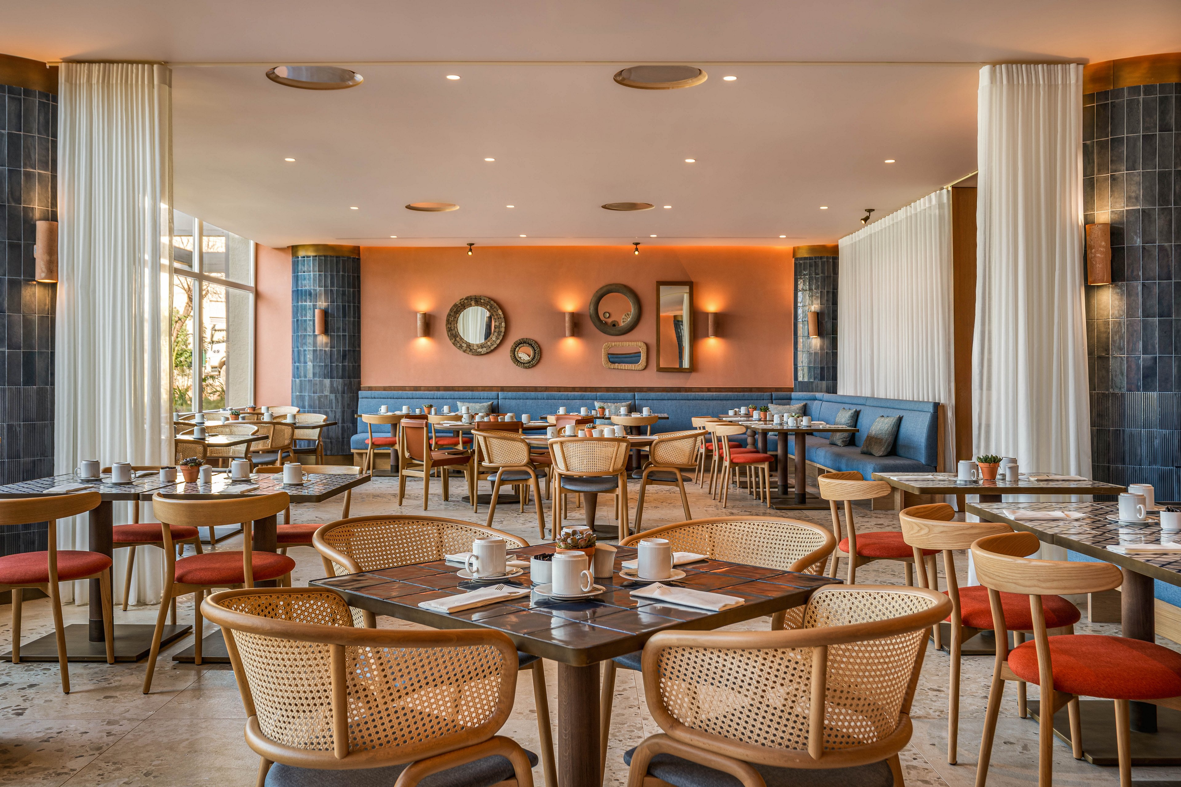 A contemporary restaurant dining room features neatly arranged wooden tables and woven chairs with orange seat cushions. The space is empty of people, highlighting the clean table settings with cups, plates, and cutlery. A warm coral accent wall with circular mirrors and wall sconces contrasts with dark blue tiled columns and white curtains. Large windows on the left bring in natural light, creating an inviting and relaxed atmosphere.