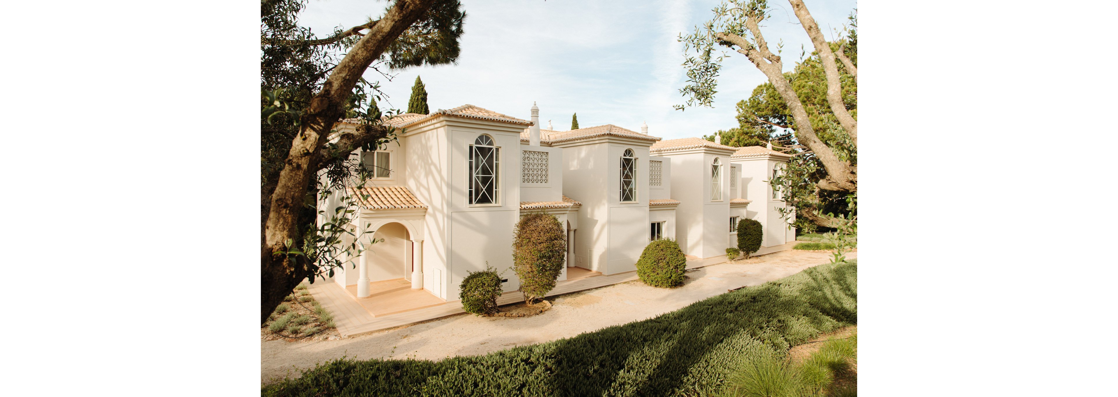 Row of white stucco villas with tiled roofs stands along a dirt path in a landscaped garden. Tall trees frame the scene, casting soft shadows on the buildings and ground. The architecture features arched entrances, decorative window grilles, and neatly trimmed bushes. Warm daylight and neutral tones create a calm, inviting residential atmosphere.