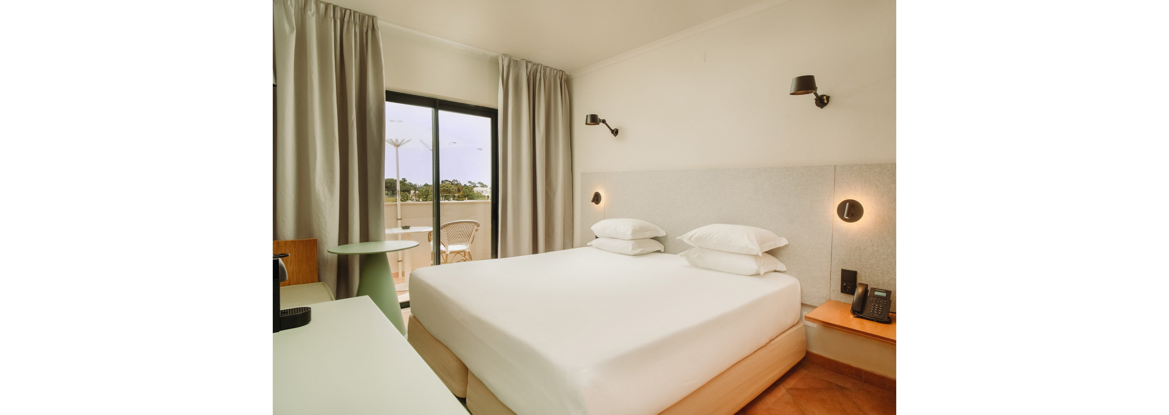 A contemporary hotel room features a large double bed with crisp white linens and two stacked pillows. Neutral walls and light curtains frame a sliding glass door that opens to a balcony with outdoor chairs and a scenic view of trees. Minimalist bedside lighting, a wall-mounted telephone, and a small desk area complete the clean, modern design. Warm flooring tones contrast with the soft, muted color palette, creating a calm and inviting atmosphere.