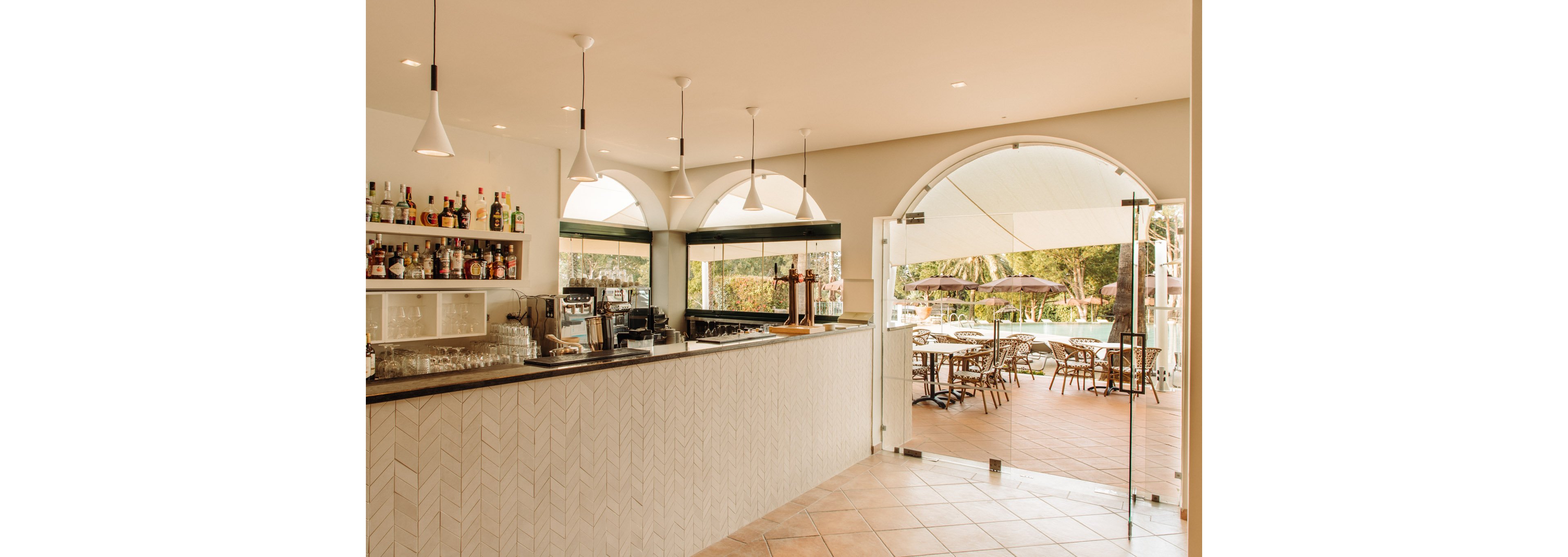 Indoor bar area with a long counter and shelves of bottles leads out to a sunny outdoor terrace. Pendant lights hang above the bar, and glassware and coffee machines are neatly arranged on the countertop. Through the open glass doors, tables and chairs are visible under umbrellas, surrounded by greenery. The warm lighting and neutral tones create a relaxed vacation atmosphere.