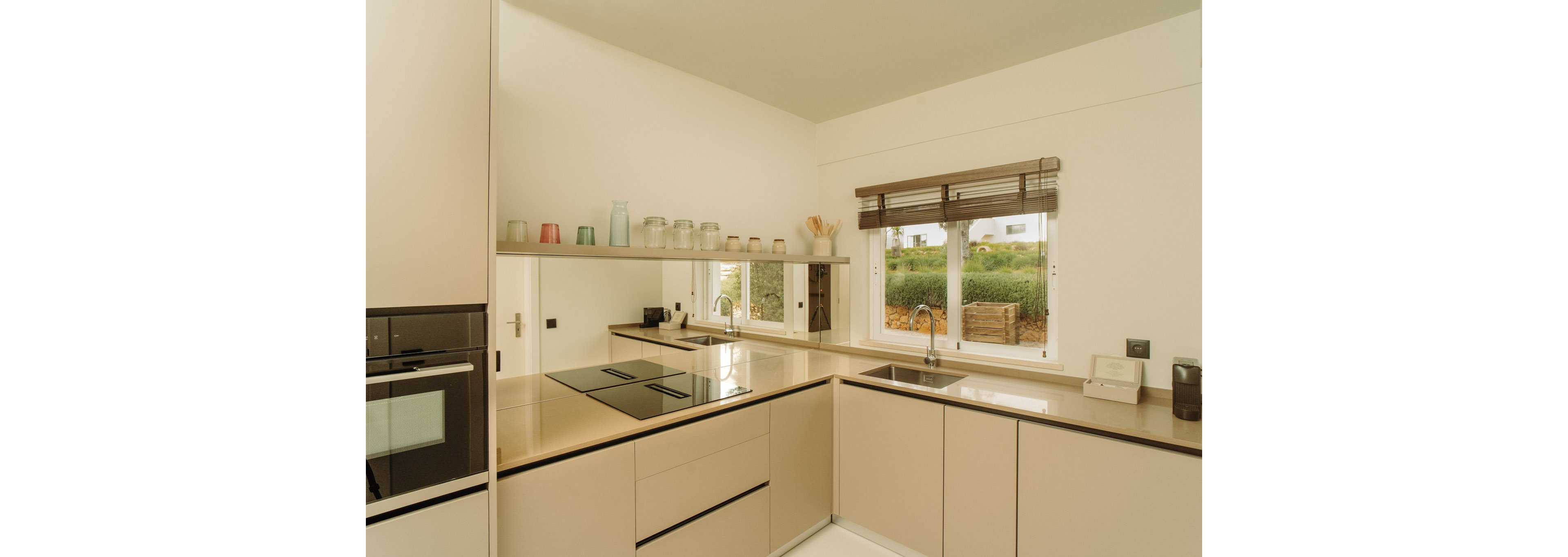 Contemporary beige kitchen interior featuring sleek cabinetry and an integrated cooktop. A long mirror backsplash reflects the room and open shelving with assorted jars and containers. Natural light enters through a double window with wooden blinds, revealing greenery and houses outside. The overall design is minimal, clean, and warm-toned.