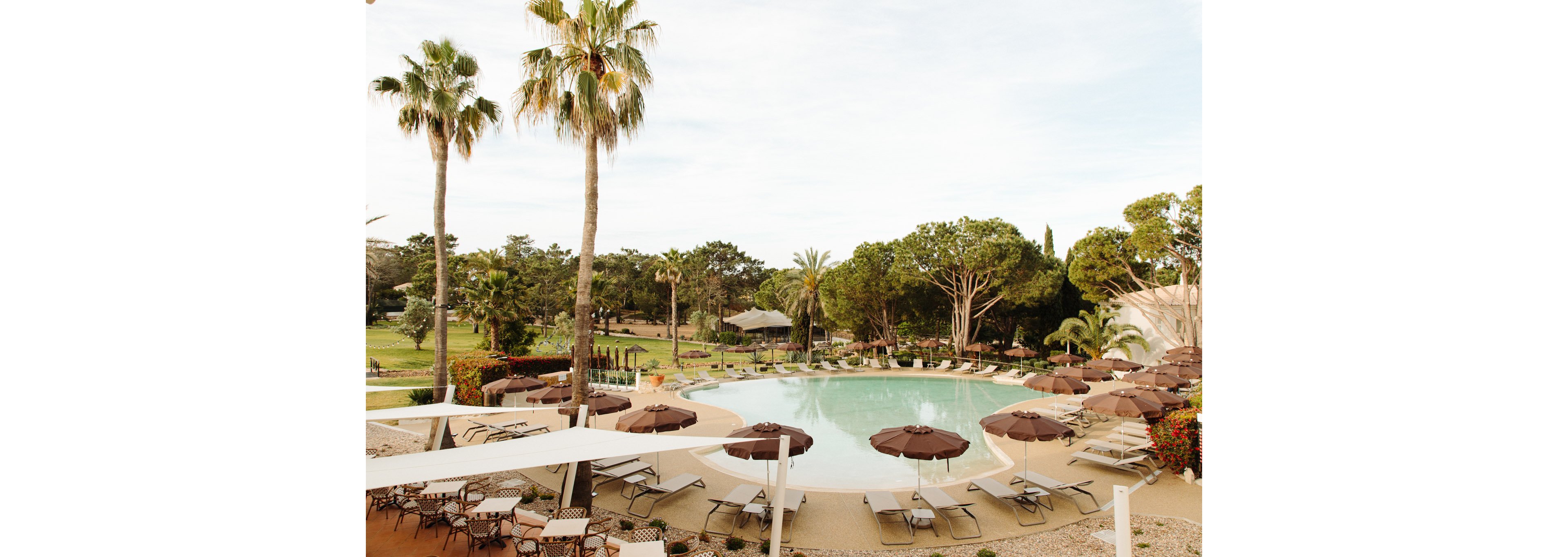 A large resort swimming pool is encircled by sun loungers and brown umbrellas in a landscaped outdoor area. Tall palm trees and lush green foliage frame the pool, creating a tropical vacation atmosphere. Tables and chairs are arranged in the foreground, suggesting an adjacent café or relaxation space. The scene is captured under soft daylight with a calm, inviting mood.