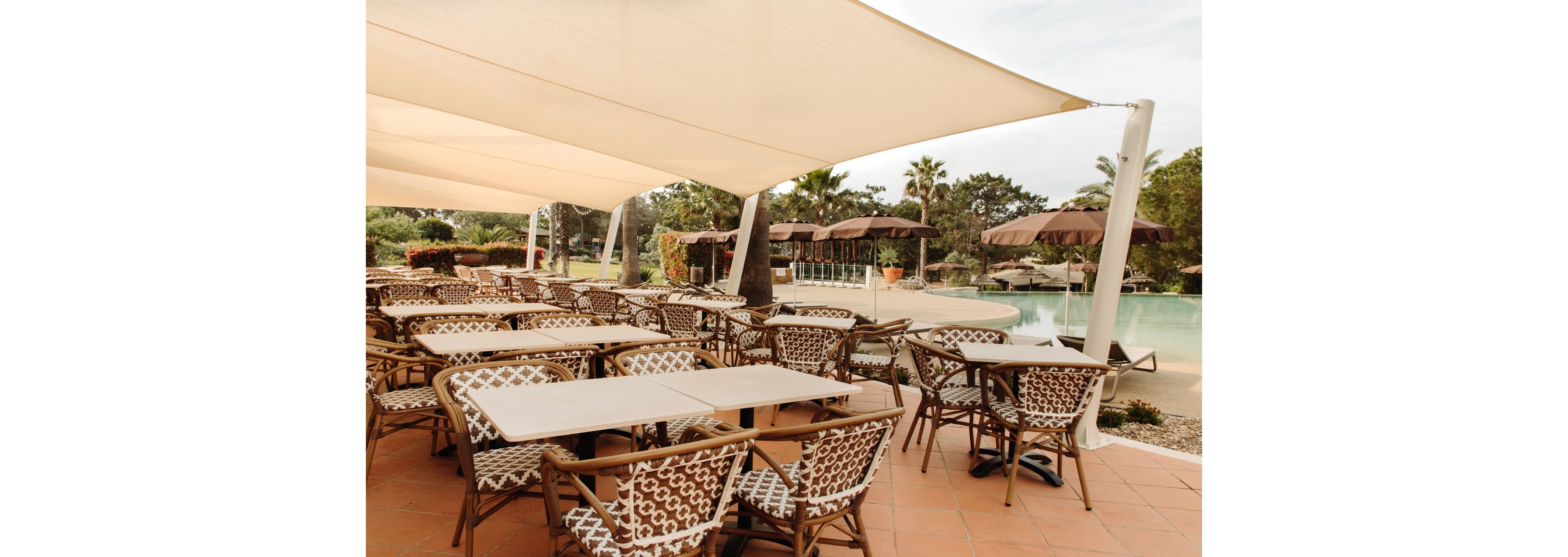 An outdoor dining terrace features multiple tables and patterned wicker chairs arranged neatly under large white shade sails. The setting overlooks a resort-style swimming pool with clear water and surrounding greenery. Brown umbrellas and palm trees enhance the relaxed vacation atmosphere. The scene is empty of people, suggesting a tranquil moment before guests arrive.