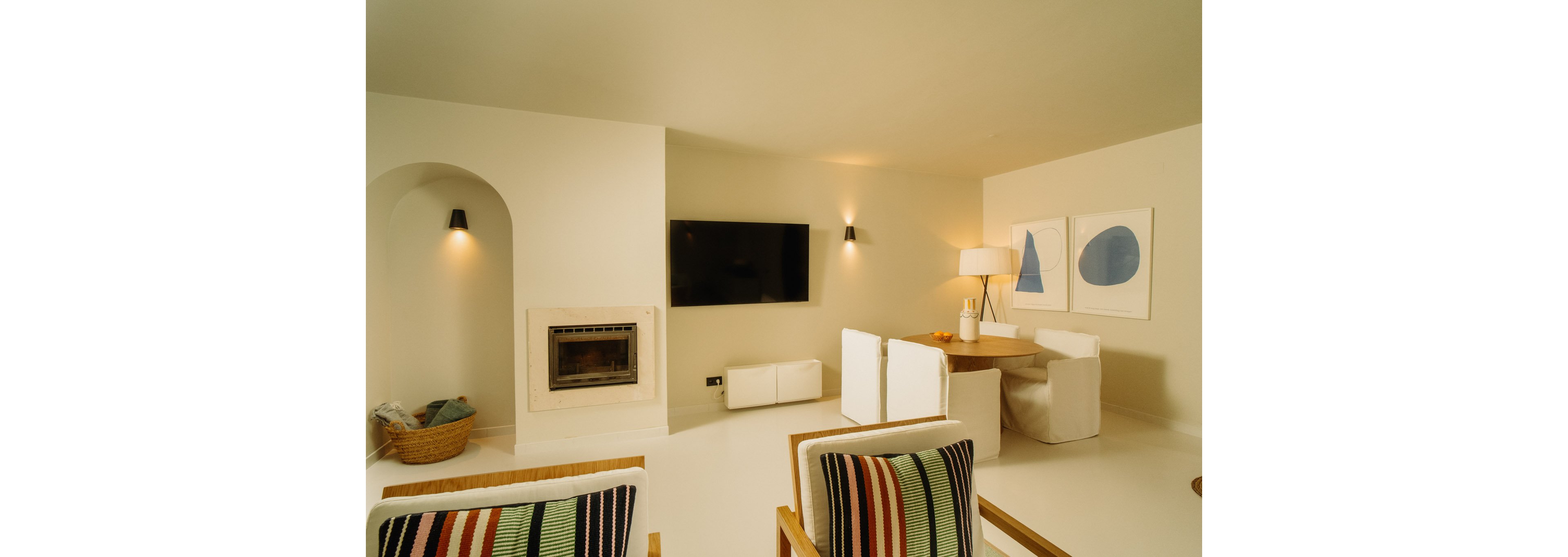 A contemporary living room features a wall-mounted flat screen television above a light-colored console and a built-in fireplace. Two armchairs with striped cushions face a round wooden table surrounded by white upholstered chairs. Soft wall sconces illuminate the neutral walls, while abstract artwork and a floor lamp add visual interest. A woven basket with blankets sits near an arched alcove, enhancing the cozy, minimalist atmosphere.