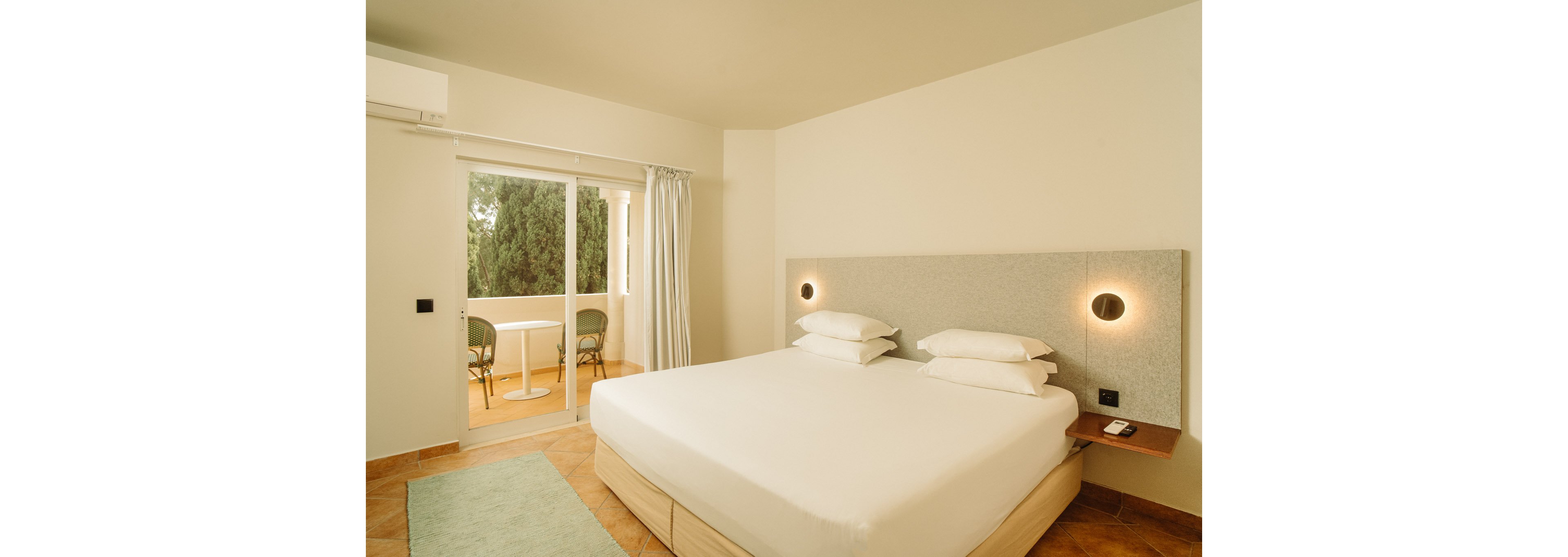 A modern hotel bedroom features a large bed with crisp white linens and multiple pillows against a light upholstered headboard. Two circular wall lights provide soft, warm illumination on either side of the bed. A sliding glass door opens to a small balcony with two chairs and a table, overlooking greenery outside. The room is decorated in neutral tones with tiled flooring and a simple rug, creating a clean and minimalist atmosphere.