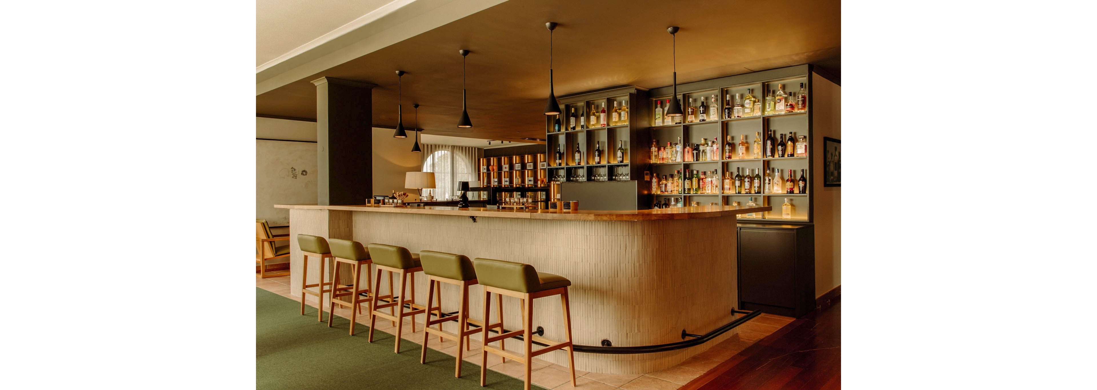 A modern bar interior features a long light-colored counter with several green upholstered bar stools. Behind the bar, tall shelves display numerous bottles of liquor and spirits in an organized grid. Pendant lights hang from the ceiling, casting warm, ambient lighting over the space. The overall design feels minimalist and sophisticated, with muted tones and clean lines.
