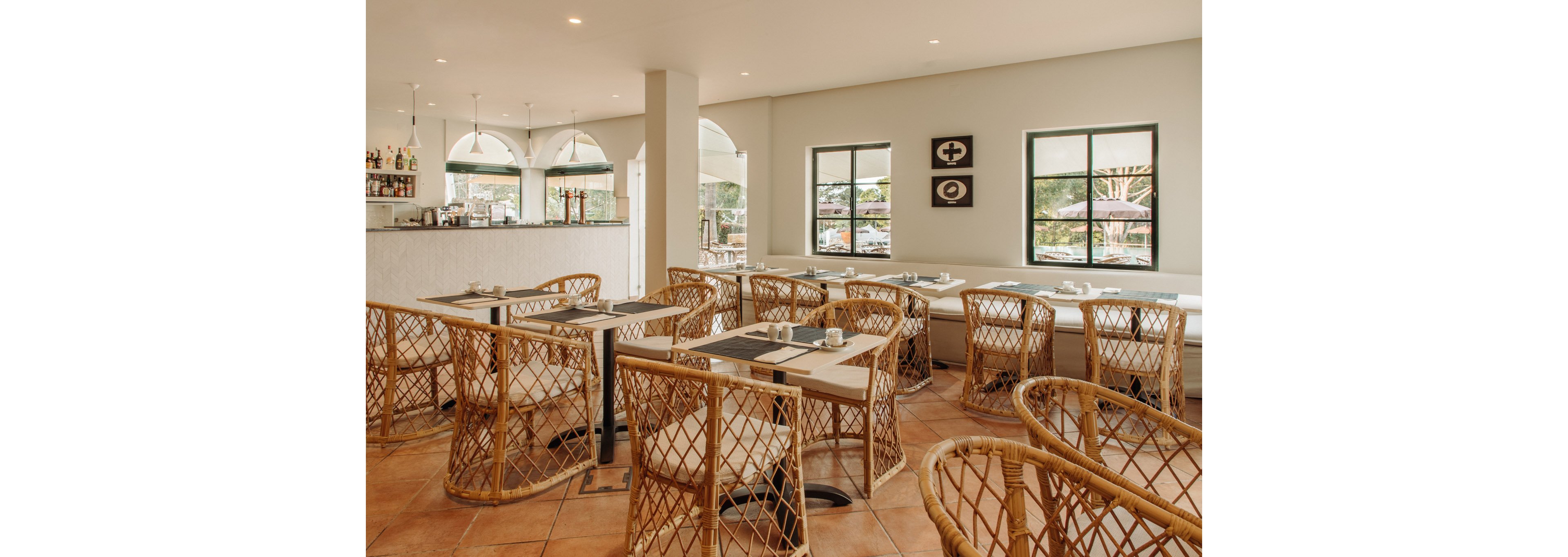 A contemporary cafe dining area features light wooden tables paired with woven wicker chairs. The space is bright and airy, with large windows and arched openings letting in natural light and views of the outdoors. A minimalist white bar counter with bottles and glassware is visible in the background. Warm tiled flooring and neutral decor create a relaxed, inviting atmosphere.