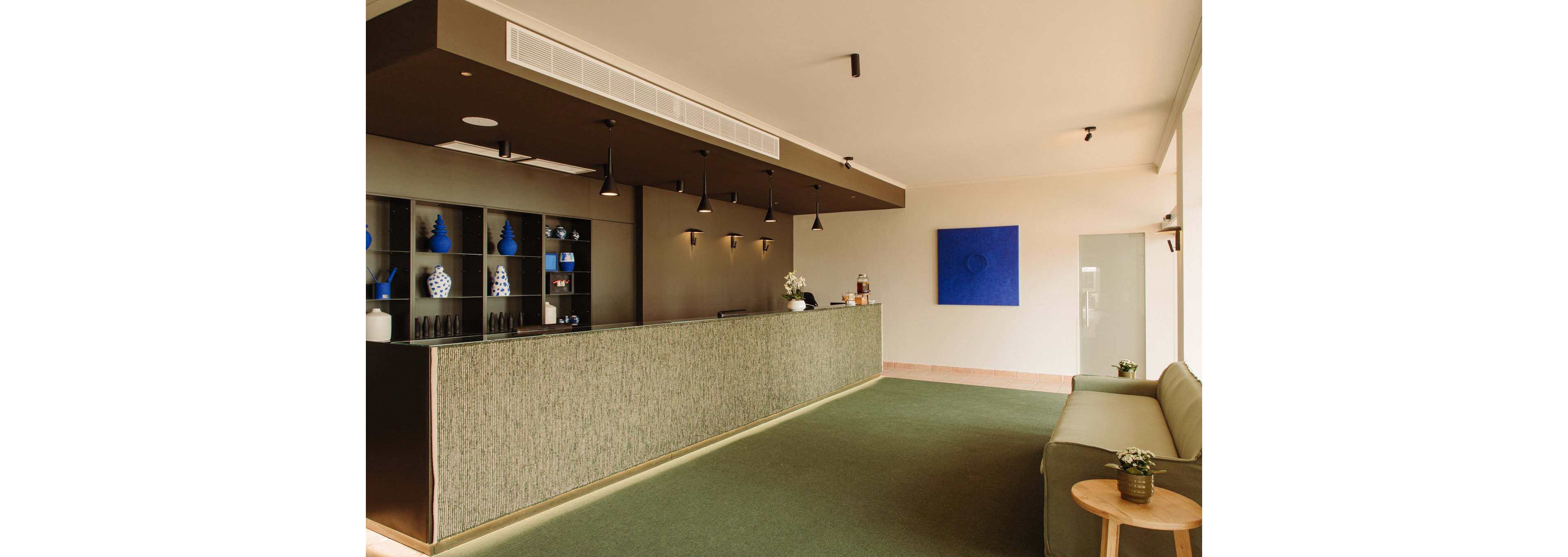 A contemporary hotel reception area features a long front desk with textured paneling and built-in shelving displaying decorative vases. Warm pendant lights hang above the counter, creating a welcoming atmosphere. A simple seating area with a sofa and small round table sits to the right, complemented by a potted plant. The room is finished with a green carpet, neutral walls, and a bold blue abstract artwork adding a pop of color.
