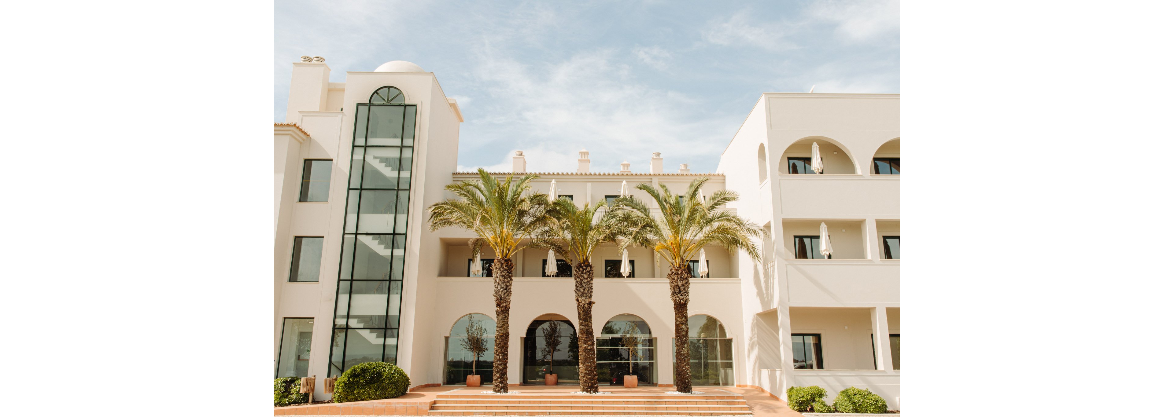 A light-colored resort building stands under a clear sky, featuring arched windows and balconies. Three tall palm trees line the front terrace, adding a tropical feel to the architecture. The structure includes a modern glass elevator shaft on the left side and multiple floors with outdoor seating areas. Warm sunlight enhances the soft, neutral tones of the facade and surrounding greenery.