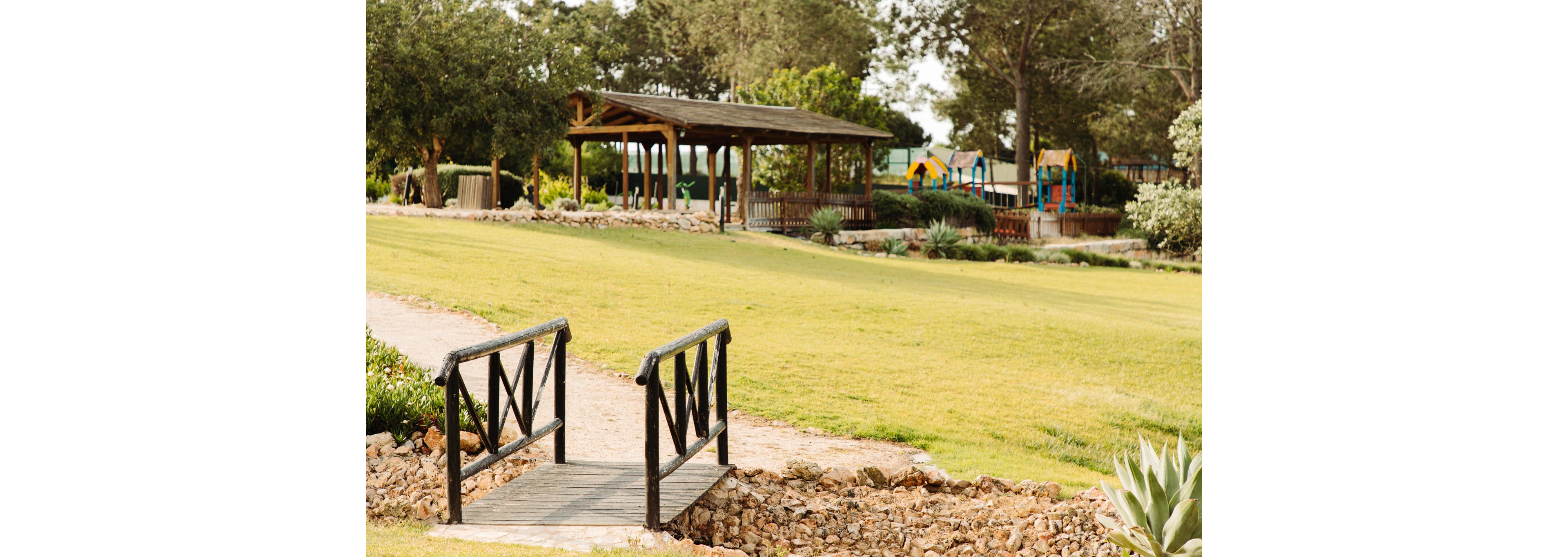 A small wooden footbridge crosses a rocky strip in a wide grassy park. In the background, a wooden pavilion with a sloped roof stands among trees and shrubs. Colorful playground structures are visible further back, adding a family-friendly feel to the scene. The overall atmosphere is calm and bright, with warm sunlight and well-maintained greenery.