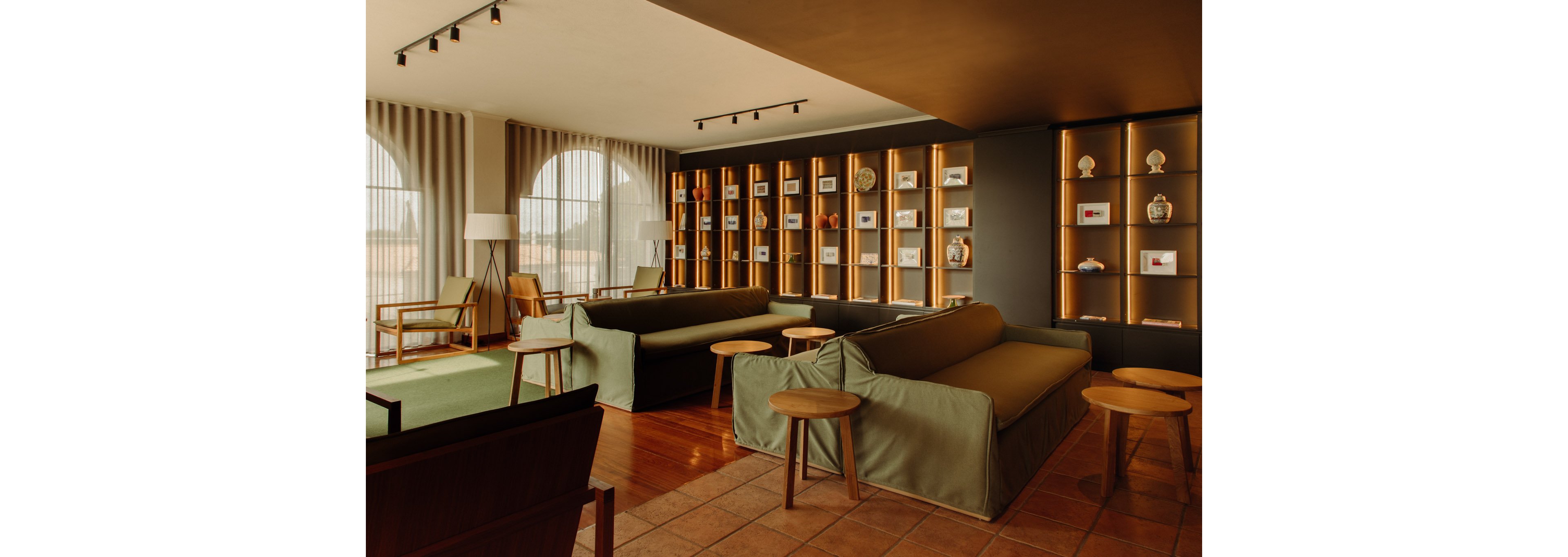 A spacious lounge features modern sofas and wooden stools arranged on a tiled floor. Large arched windows with draped curtains allow soft natural light into the room. Along one wall, illuminated shelving displays decorative objects and framed items, adding visual interest. The overall atmosphere is warm and inviting, with earthy tones and minimalist contemporary decor.