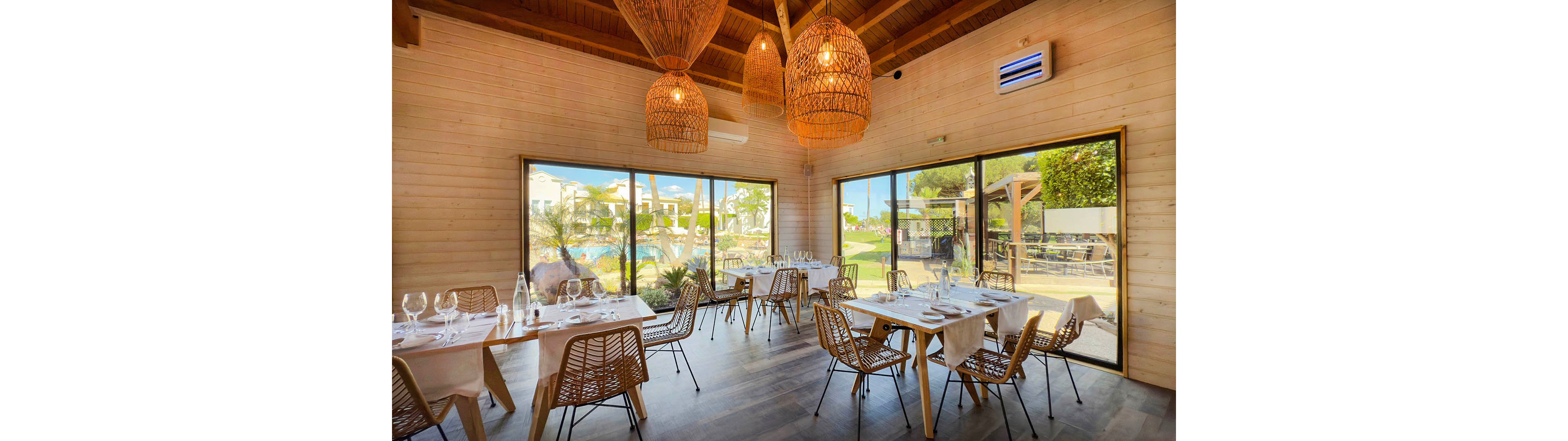 Contemporary restaurant interior featuring wooden walls, wicker chairs, and neatly set dining tables. Large floor-to-ceiling windows open onto a sunny outdoor pool and garden area. Warm pendant lights hang from the wooden ceiling, creating a cozy yet airy atmosphere. The space appears ready for guests, with glassware and table linens arranged for service.