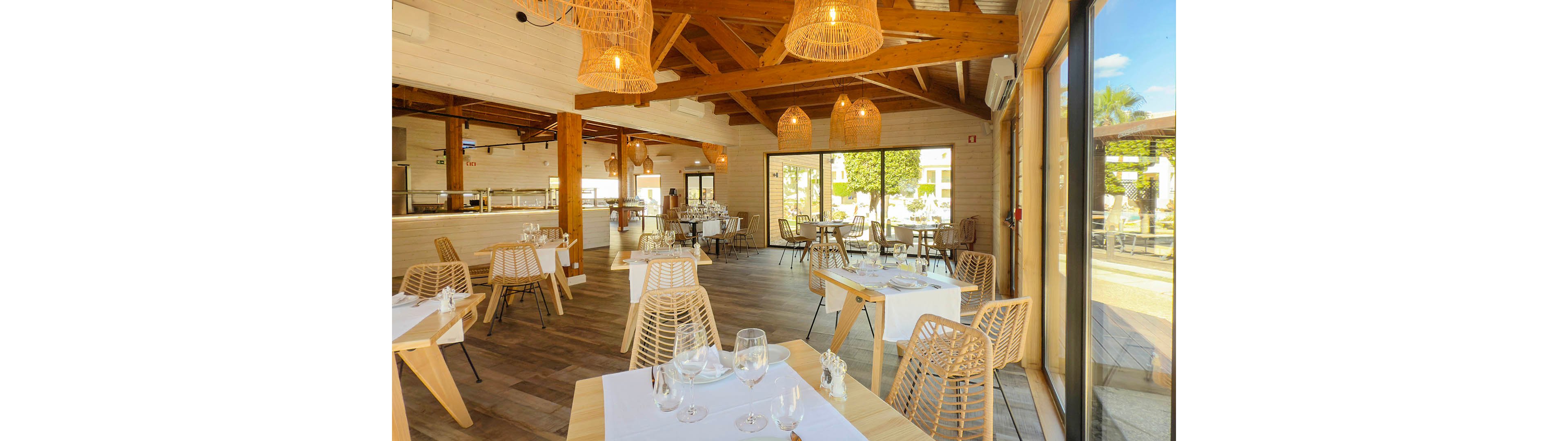 Contemporary restaurant interior featuring light wooden furniture and exposed beam ceilings. Sunlight streams through large glass windows, illuminating neatly set tables with white tablecloths and glassware. Woven pendant lights hang from the ceiling, adding warmth and texture to the space. The overall atmosphere is airy, inviting, and relaxed, suitable for casual or upscale dining.