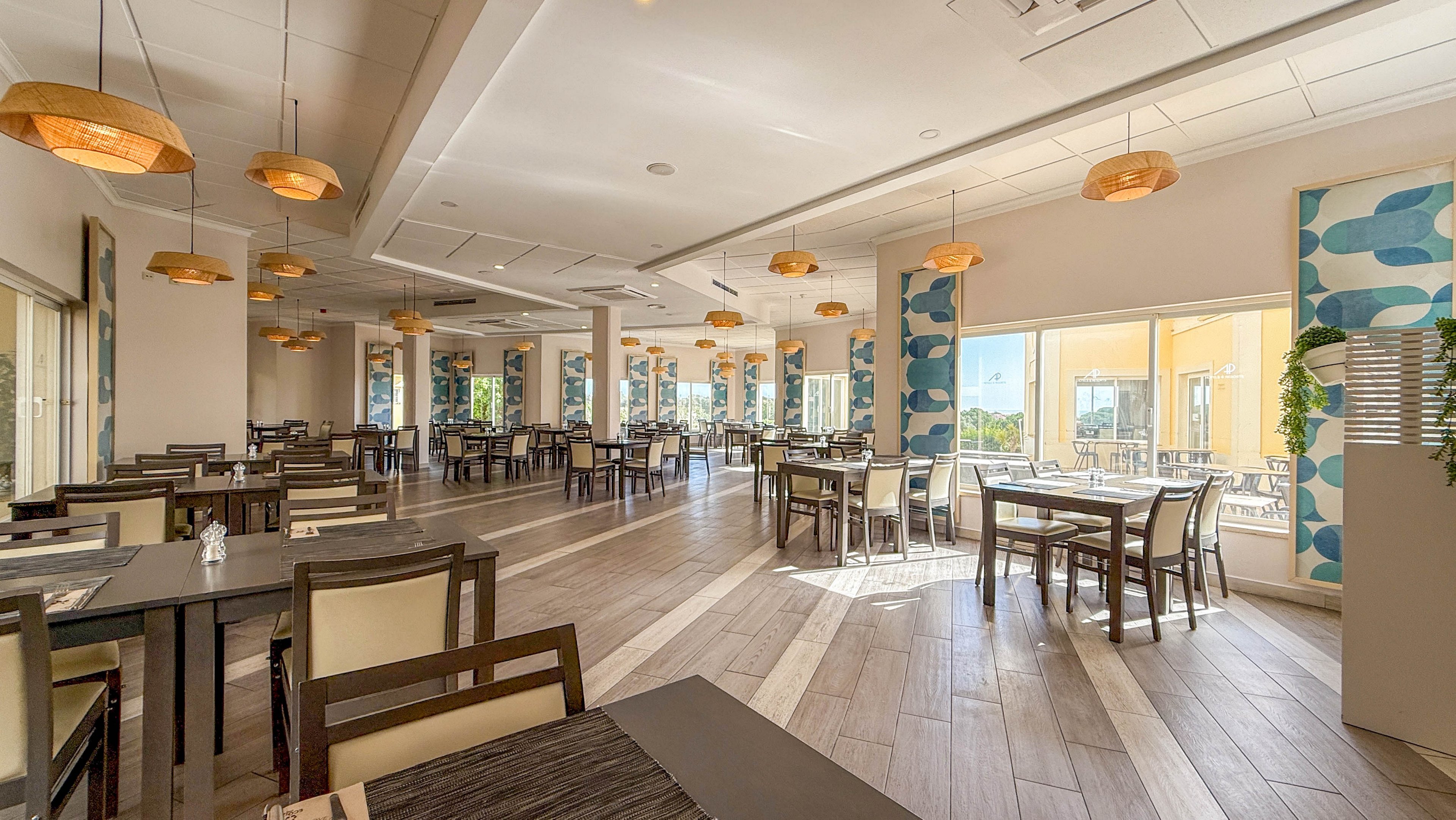 A large, contemporary restaurant dining area features neatly arranged tables and chairs, illuminated by ample natural light from wide windows. The setting is clean and inviting, with wooden flooring, modern pendant lights, and decorative blue patterned wall panels. The space appears empty, emphasizing its calm and airy atmosphere. Outdoor seating is visible through the glass, enhancing the open feel.