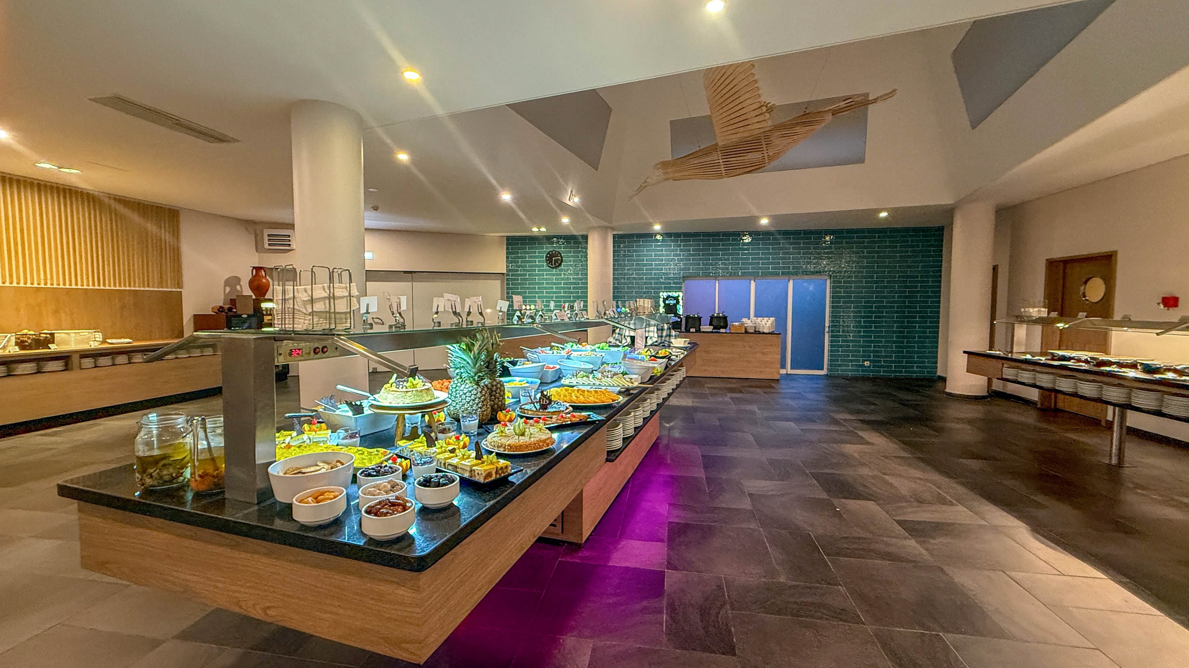 A spacious, contemporary buffet area features a central island filled with a variety of fresh salads, fruits, and appetizers. The setting is a well-lit dining room with tiled floors, teal accent wall, and minimalist decor. The buffet table is illuminated with purple lighting underneath, and glass containers and serving dishes are neatly arranged. No visible text or numbers are present in the image. The atmosphere is clean, inviting, and organized for self-service dining.