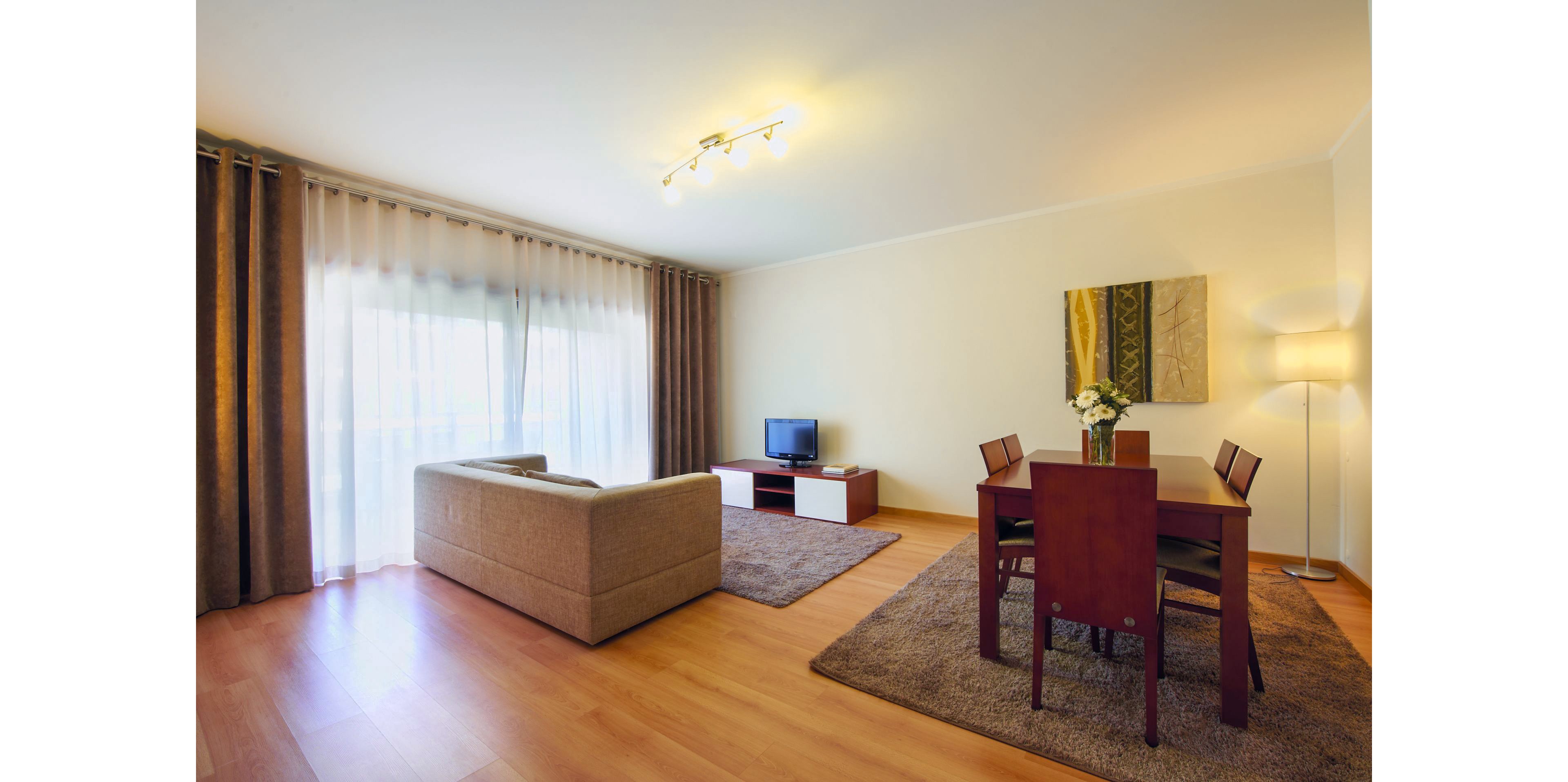A contemporary living room features a beige sofa facing a small television on a low white and wood media unit. Floor-to-ceiling curtains cover a large window, allowing soft natural light into the space. A wooden dining table with four chairs sits on a brown rug, decorated with a vase of white flowers. Warm wall lighting and a standing lamp create a cozy, inviting atmosphere with neutral tones and clean lines.