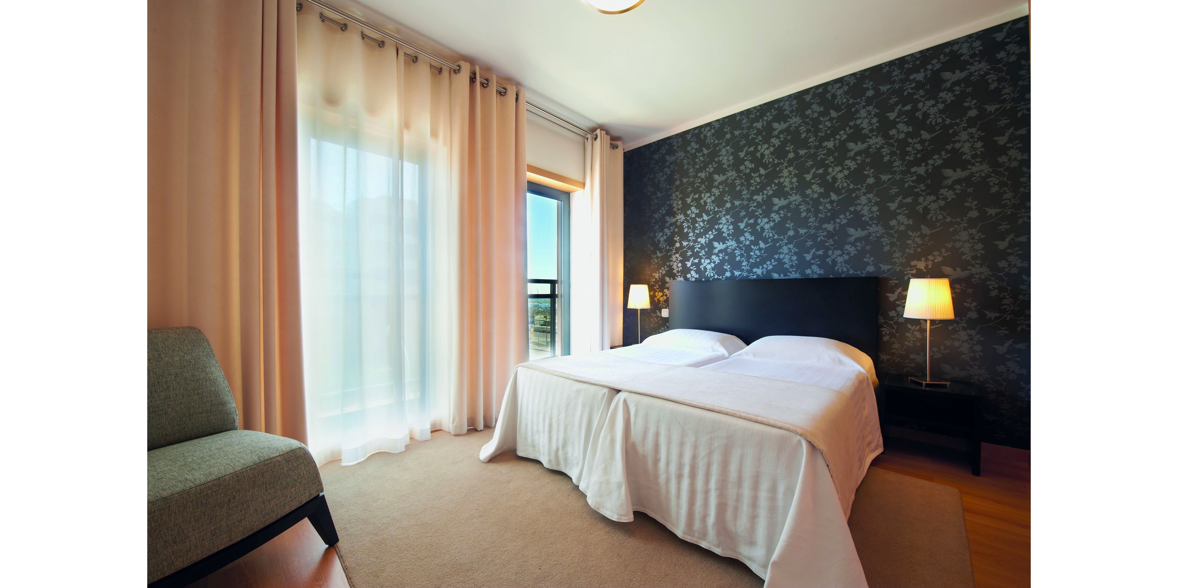 Contemporary bedroom interior featuring two neatly made twin beds with white linens and a dark upholstered headboard. The room includes soft beige curtains covering a large window and glass door leading to a balcony with an outdoor view. Warm lighting from two bedside table lamps highlights the patterned dark accent wall behind the bed. A cushioned armchair and light-colored carpet add comfort and a minimalist, modern style to the space.
