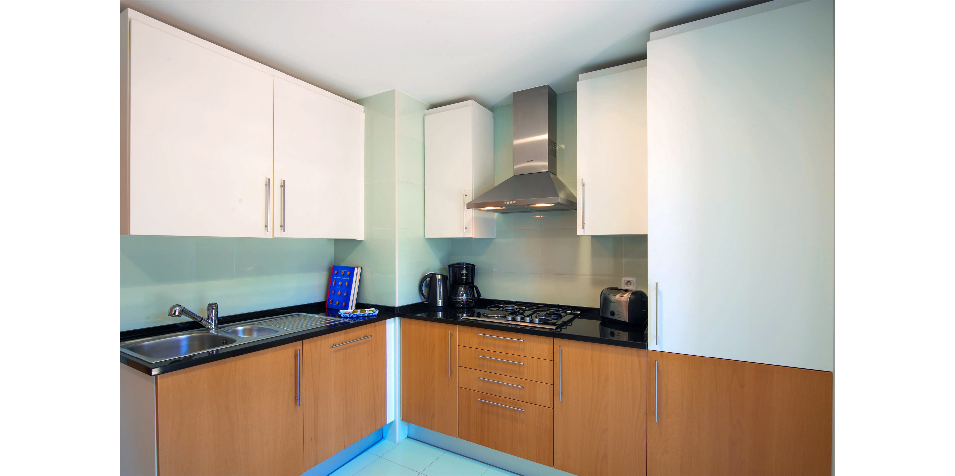 A modern corner kitchen features light wood lower cabinets and white upper cabinets with sleek metal handles. A stainless steel range hood sits above a gas cooktop, flanked by small appliances including a kettle, coffee maker, and toaster. A double sink with a chrome faucet is installed on the black countertop opposite a light-colored tiled floor. The overall design is clean and contemporary, emphasizing functionality in a compact space.