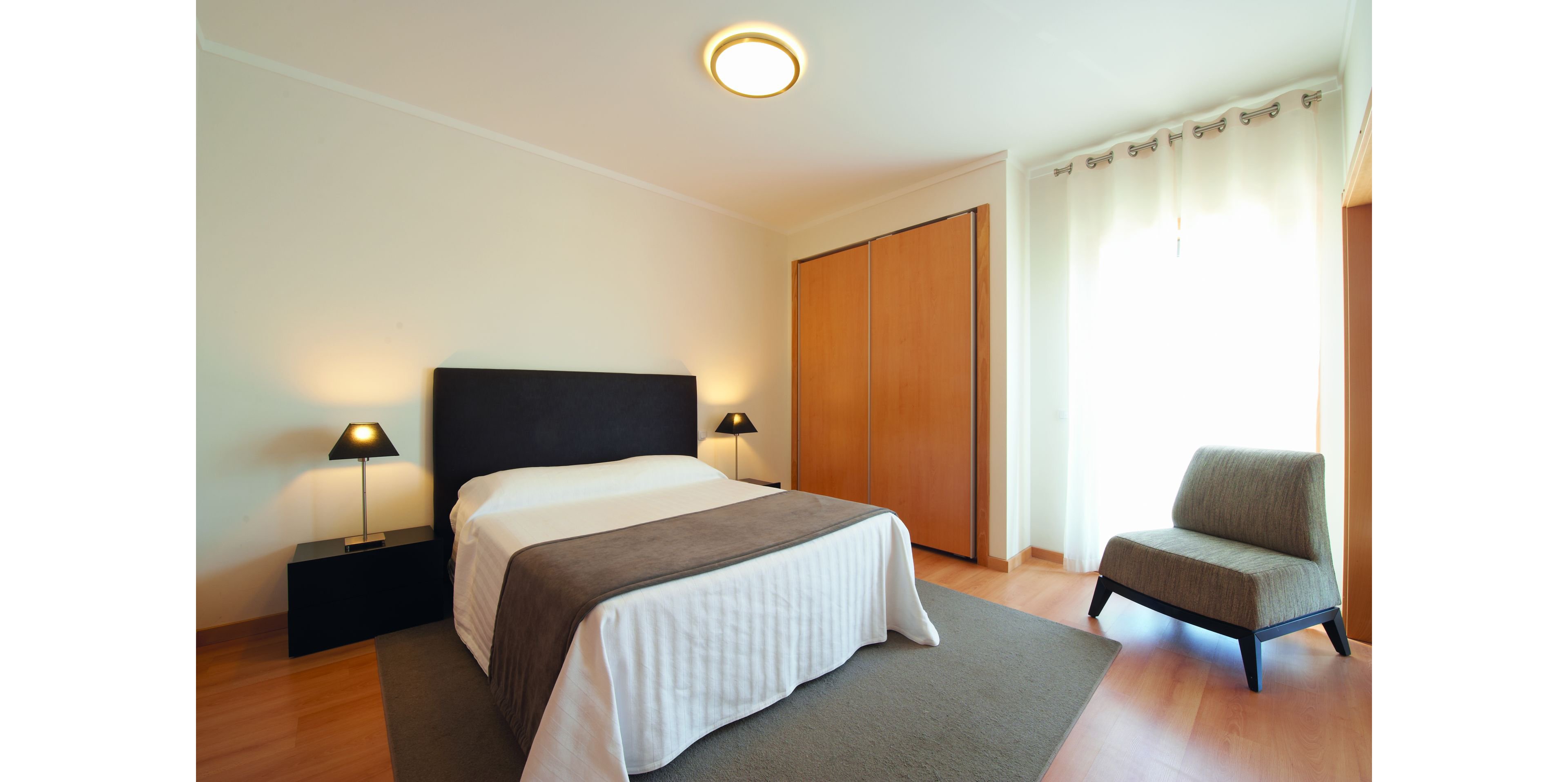 A contemporary bedroom features a double bed with a dark headboard, white bedding, and a gray throw blanket. Two bedside tables with matching lamps flank the bed, adding symmetry and soft lighting. A large wooden wardrobe stands against the wall, while a cushioned armchair sits near sheer curtains that let in bright natural light. The room has light wood flooring and a neutral color palette, creating a clean and minimalist atmosphere.