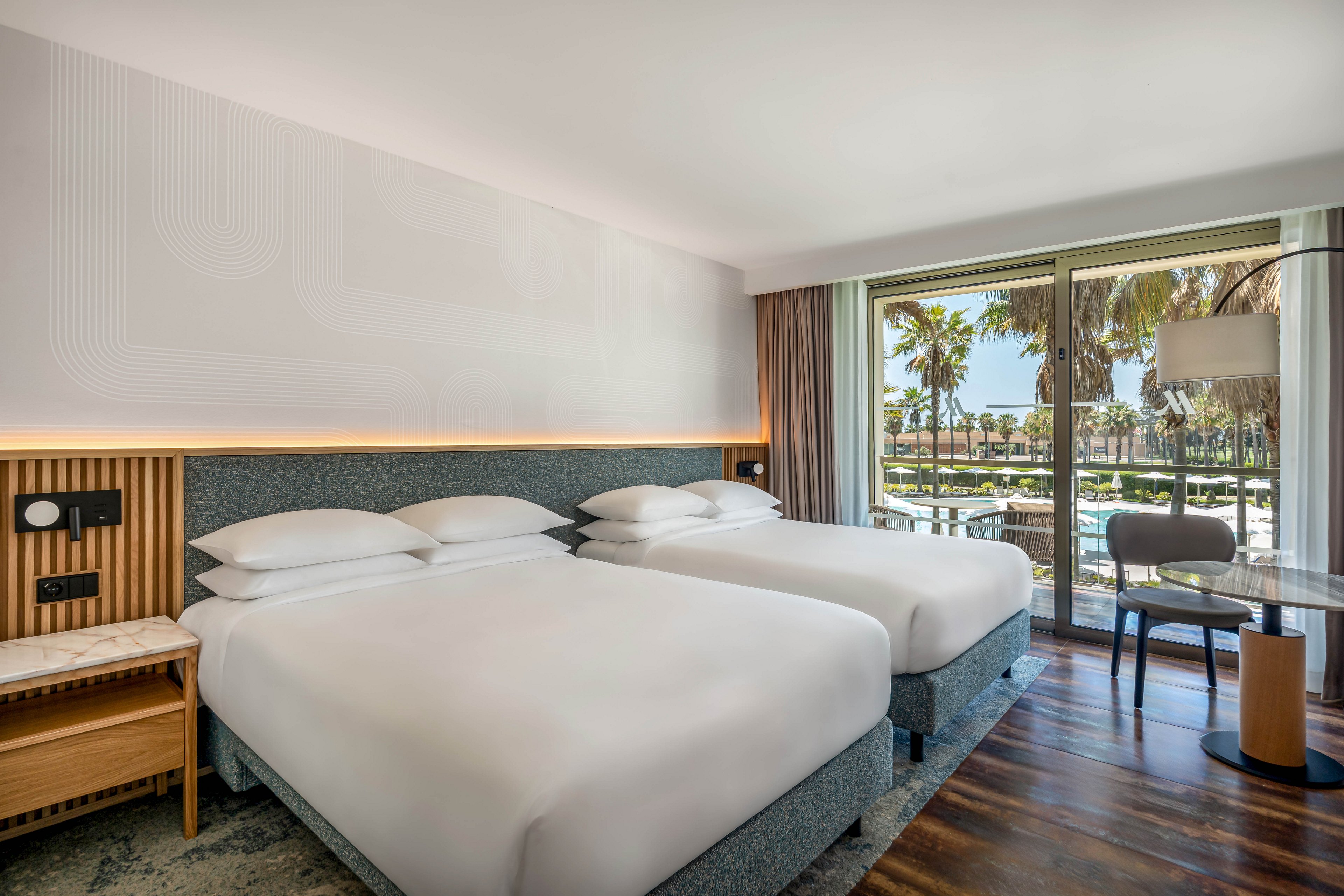 A contemporary hotel guest room features two neatly made double beds with white linens and a minimalist headboard. The space includes a wooden nightstand, wall-mounted reading lights, and a small seating area with a round glass table and chair. Large floor-to-ceiling windows open to a balcony overlooking palm trees and a sunny outdoor area. Natural light and neutral tones create a bright, calm, and inviting atmosphere.