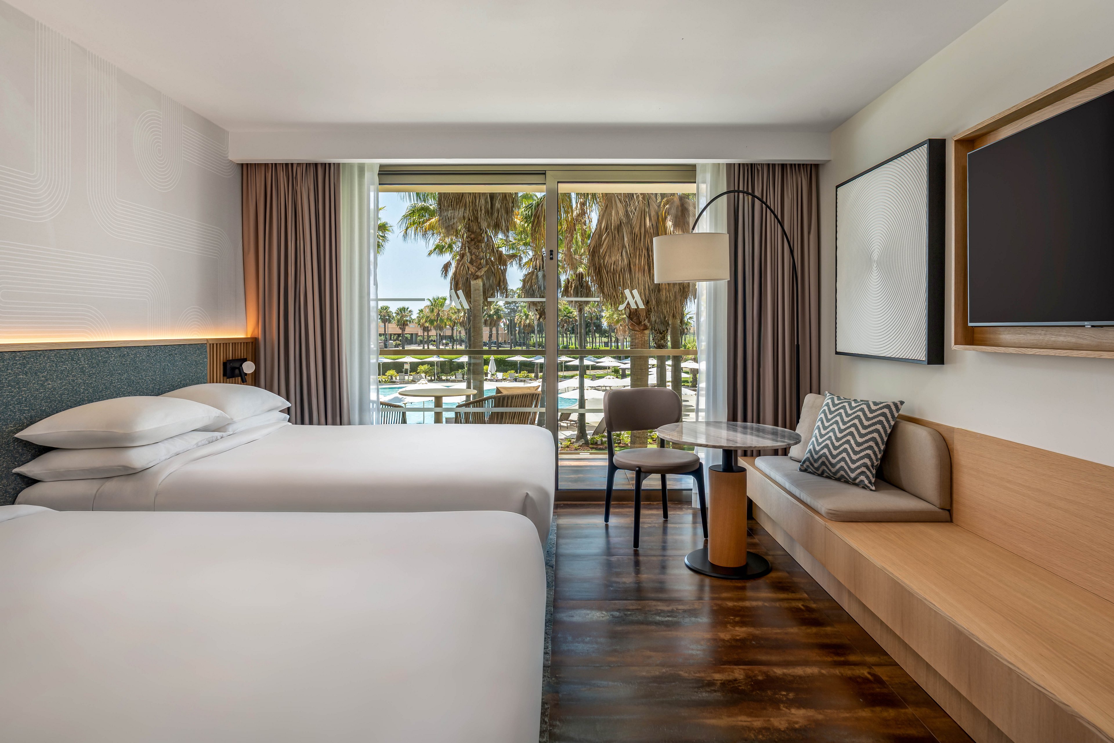 A contemporary hotel room features two neatly made twin beds with white linens and a minimalist headboard. The space includes a built-in wooden bench, a small round table, and a floor lamp beside a large wall-mounted television. Floor-to-ceiling curtains frame sliding glass doors that open to a balcony overlooking palm trees and a waterfront area. Neutral tones and clean lines create a calm, modern atmosphere ideal for relaxation.