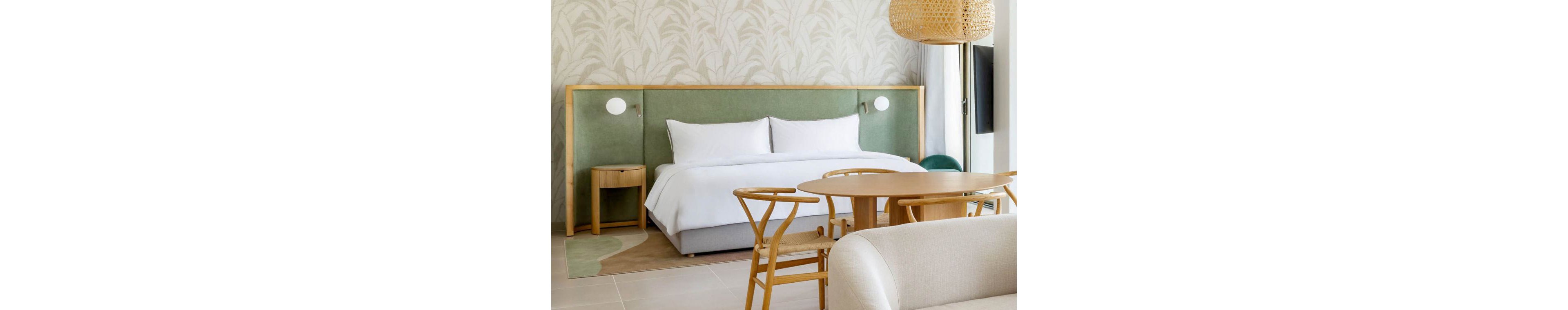 A contemporary hotel bedroom features a large neatly made bed with white linens and a light green upholstered headboard. The room includes a small wooden bedside table, a round wooden dining table, and matching chairs. Soft neutral tones, natural wood accents, and a woven pendant light create a calm, minimalist atmosphere. A patterned wall and subtle area rug add texture while maintaining a clean, modern design.