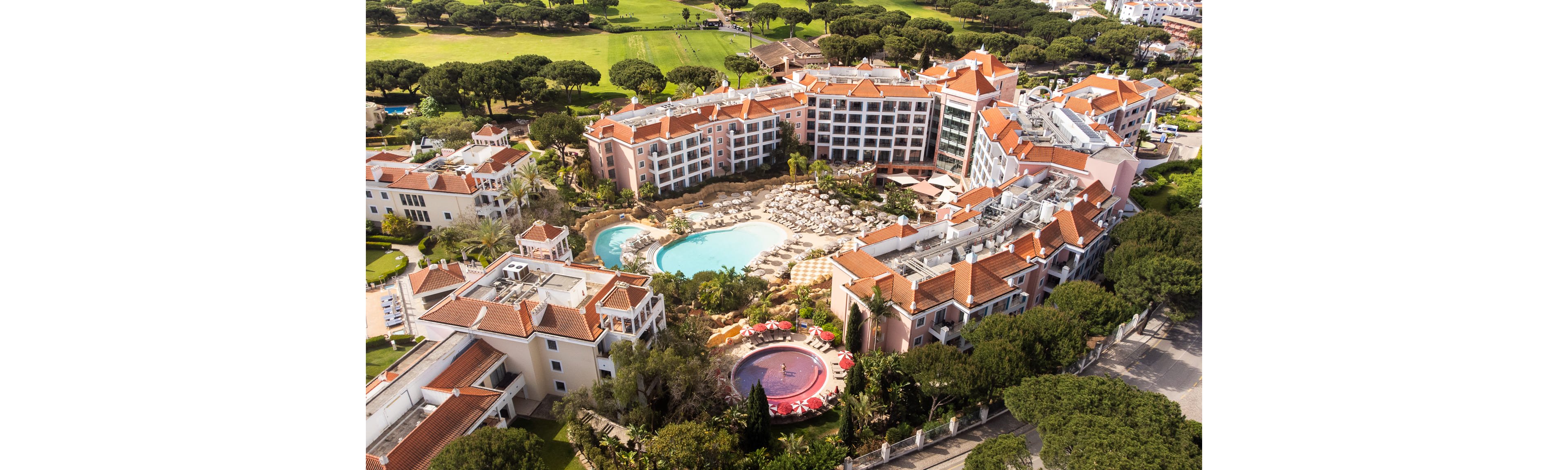 A large resort complex with multiple buildings surrounds two outdoor swimming pools, one turquoise and one circular with red loungers. The property is nestled among dense green trees and landscaped gardens, with a golf course visible in the background. Sun loungers and umbrellas line the poolside terraces, suggesting a relaxing vacation atmosphere. The architecture features light-colored facades and red-tiled roofs, creating a Mediterranean-style setting.
