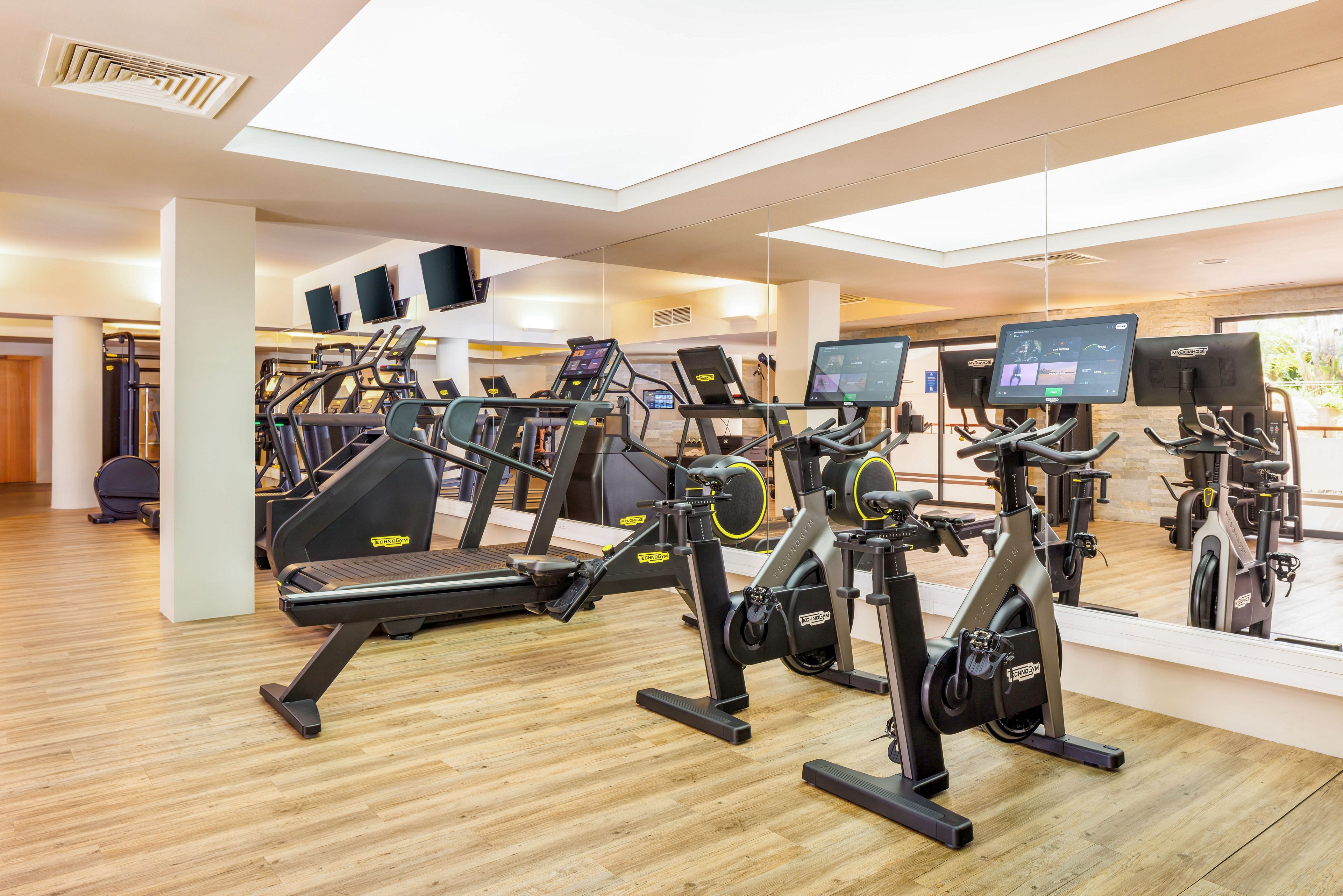 A bright, modern gym features several stationary bikes and treadmills arranged in front of a large wall mirror. Digital screens are mounted on the cardio machines, adding a high-tech feel to the space. The room has light wooden flooring, neutral walls, and recessed ceiling lighting that creates an airy atmosphere. No people are present, emphasizing the clean, organized layout of the fitness equipment.