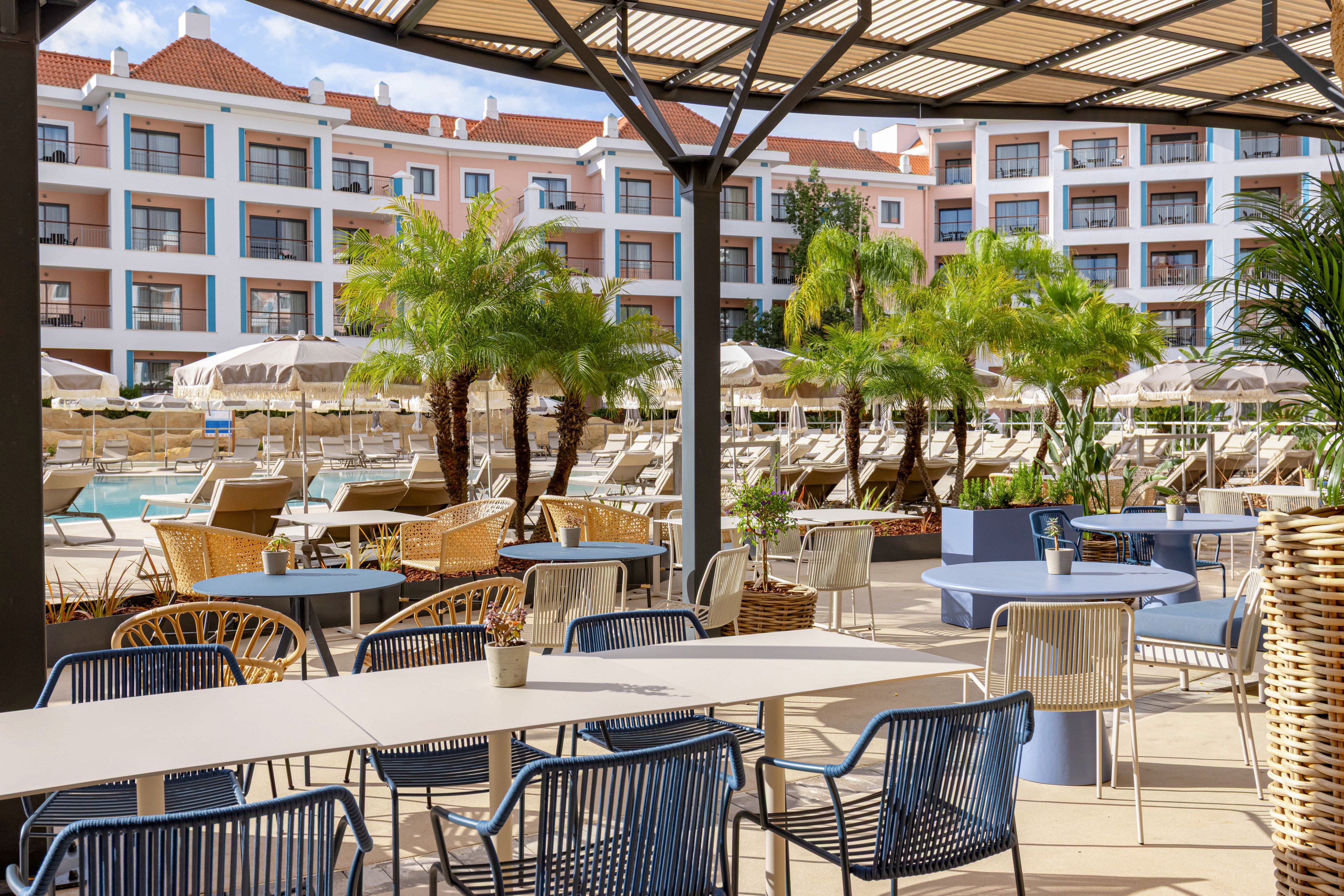 An inviting outdoor terrace with tables and chairs overlooks a resort swimming pool area. Sun loungers and umbrellas are arranged around the pool, framed by tall palm trees. In the background, a multi-story hotel building with balconies and large windows completes the scene. The atmosphere is bright and relaxed, suggesting a vacation or leisure setting in warm weather.