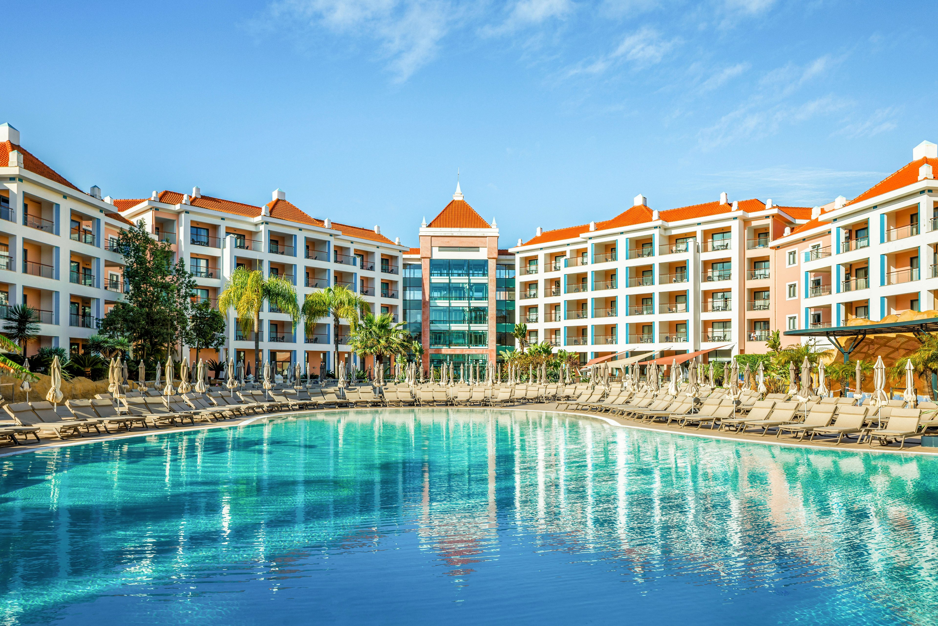 A modern resort hotel with red-tiled roofs surrounds a large, clear blue swimming pool. Rows of sun loungers and umbrellas line the poolside, suggesting a relaxing vacation setting. The multi-story buildings feature balconies overlooking the water and landscaped palm trees. The bright daylight and vivid colors create a warm, inviting holiday atmosphere.