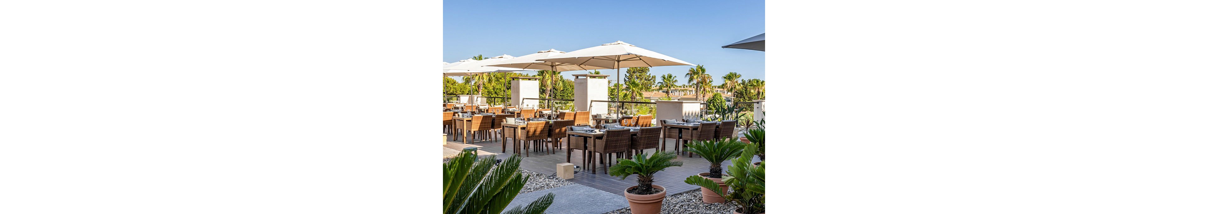 An open-air restaurant terrace features neatly arranged wooden tables and wicker chairs under large white sun umbrellas. Place settings are prepared on each table, suggesting readiness for guests in a relaxed dining environment. Potted green plants and gravel landscaping frame the seating area, adding a touch of nature. In the background, palm trees and clear blue sky create a warm, resort-like atmosphere.