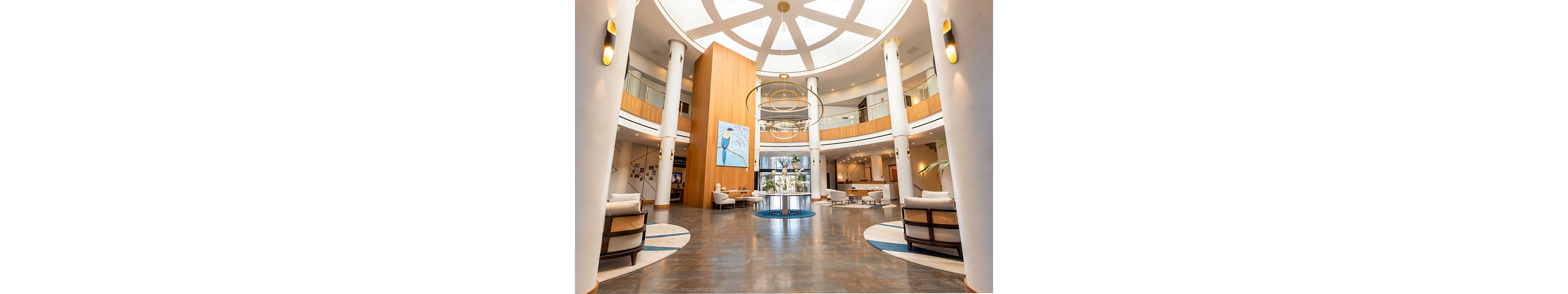 Spacious contemporary hotel lobby featuring a circular atrium and high ceiling with geometric skylight pattern. The central area includes a wooden column with modern artwork and large circular pendant lights. Comfortable seating areas with armchairs and sofas are arranged around the perimeter, complemented by warm wood tones and white columns. Natural light enters from glass doors at the far end, enhancing the bright and welcoming atmosphere.
