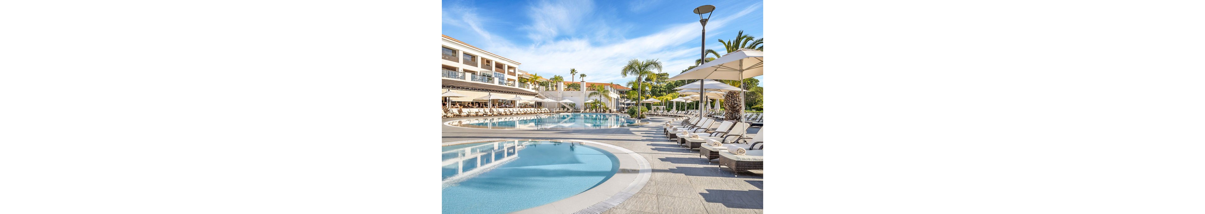 A large curved swimming pool is surrounded by rows of white sun loungers and umbrellas at a modern resort. The multi-story hotel buildings frame the pool area, featuring balconies and shaded terraces. Palm trees and manicured landscaping add a tropical feel under a bright blue sky with light clouds. The scene suggests a relaxing vacation atmosphere in a clean, upscale outdoor setting.