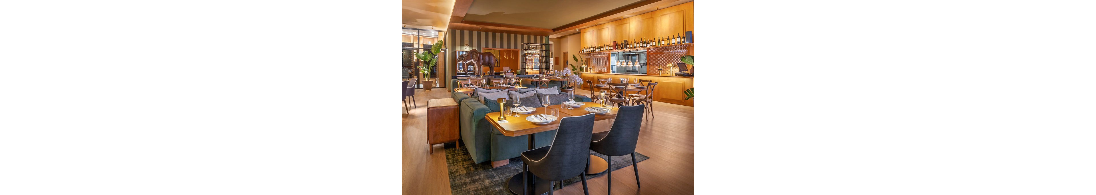 Upscale restaurant dining area featuring wooden tables, upholstered chairs, and a long teal sofa. The setting includes a warm, well-lit bar with bottles displayed on shelves and pendant lights above the counter. Decorative elements such as a large horse sculpture, plants, and striped wall panels add visual interest. Table settings with plates, glasses, and cutlery suggest readiness for guests in a cozy, modern atmosphere.