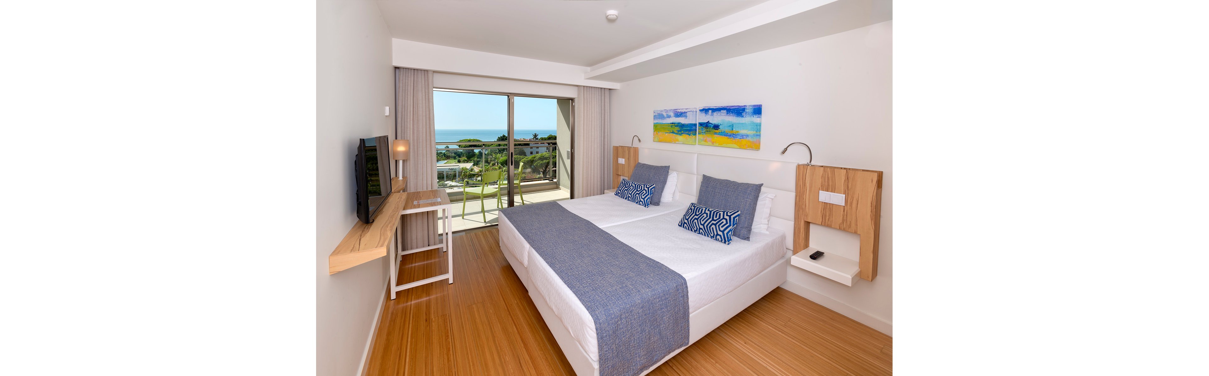 Modern hotel room featuring two neatly made single beds with blue throws and patterned pillows. A sliding glass door opens to a balcony overlooking greenery and the distant ocean. Light wood flooring and minimalist furniture create a clean, contemporary look. Colorful abstract artwork and natural daylight contribute to a fresh, relaxing atmosphere.