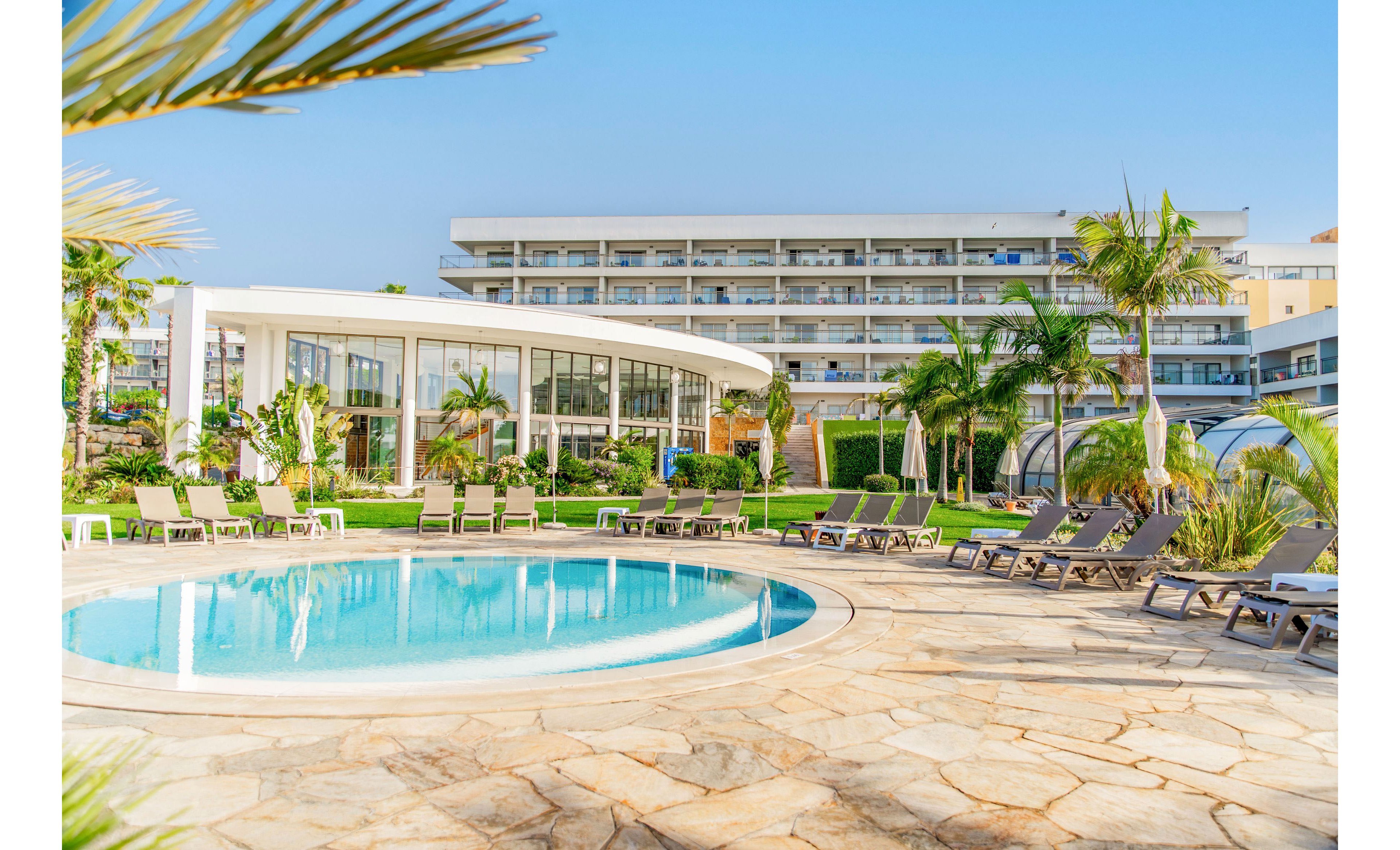 A circular outdoor swimming pool is surrounded by sun loungers and umbrellas in a landscaped resort courtyard. Palm trees and green lawns create a tropical atmosphere under a clear blue sky. Behind the pool, a modern glass-fronted building and a multi-story hotel with balconies are visible. The scene suggests a relaxing vacation destination with ample seating and open space.