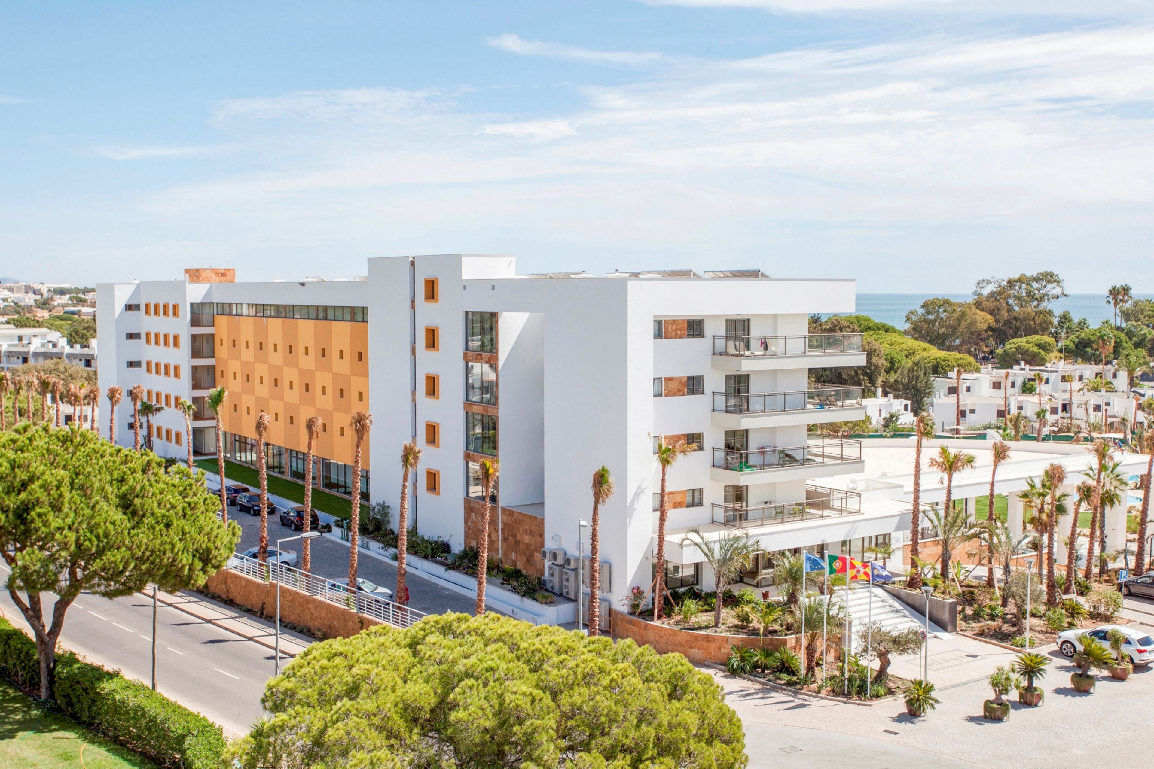 A contemporary white and ochre apartment complex stands along a sunny coastal road. The building features multiple balconies, large windows, and clean geometric lines. Palm trees and landscaped greenery surround the property, with parked cars and walkways visible in the foreground. In the distance, additional white buildings and a glimpse of the sea suggest a Mediterranean or resort-style setting.