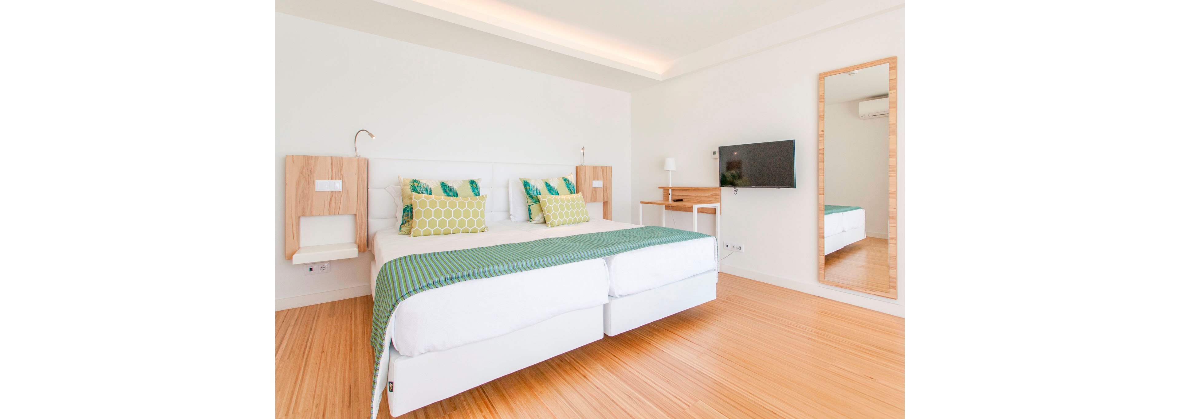 A contemporary bedroom features two white twin beds pushed together, topped with green throws and patterned pillows. The room has light wooden flooring and minimalist white walls, creating an airy, spacious feel. Built-in wooden bedside units, a wall-mounted flat-screen TV, and a small desk add functional touches. A tall framed mirror reflects the bed area, enhancing the sense of light and openness.