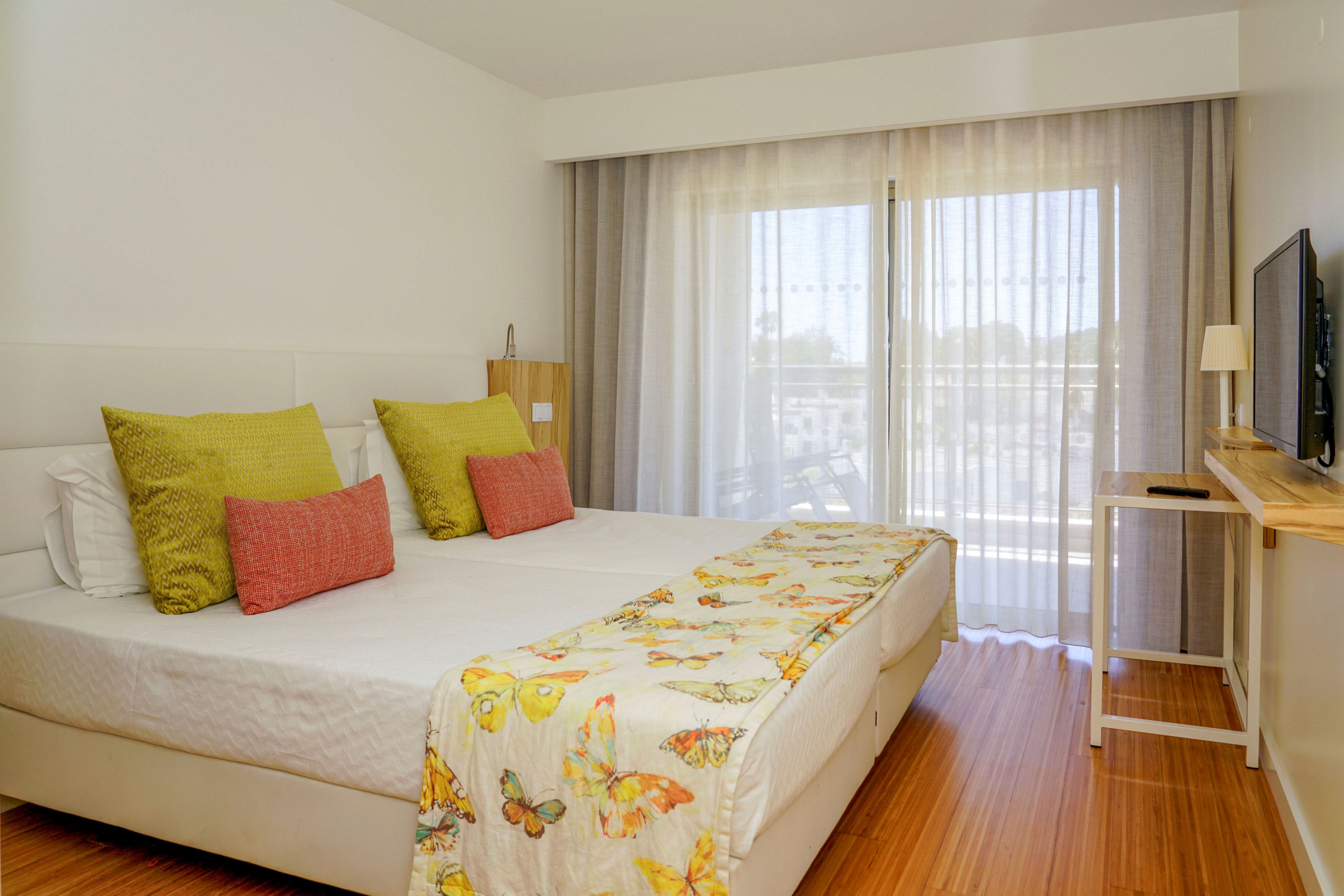 A contemporary hotel bedroom features a large bed with white linens and colorful accent pillows. A butterfly-patterned throw adds a cheerful touch to the room. Floor-to-ceiling sheer curtains cover the glass doors leading to a balcony with an outdoor view. A flat-screen television is mounted on a wooden shelf opposite the bed, complementing the clean, minimalist decor.