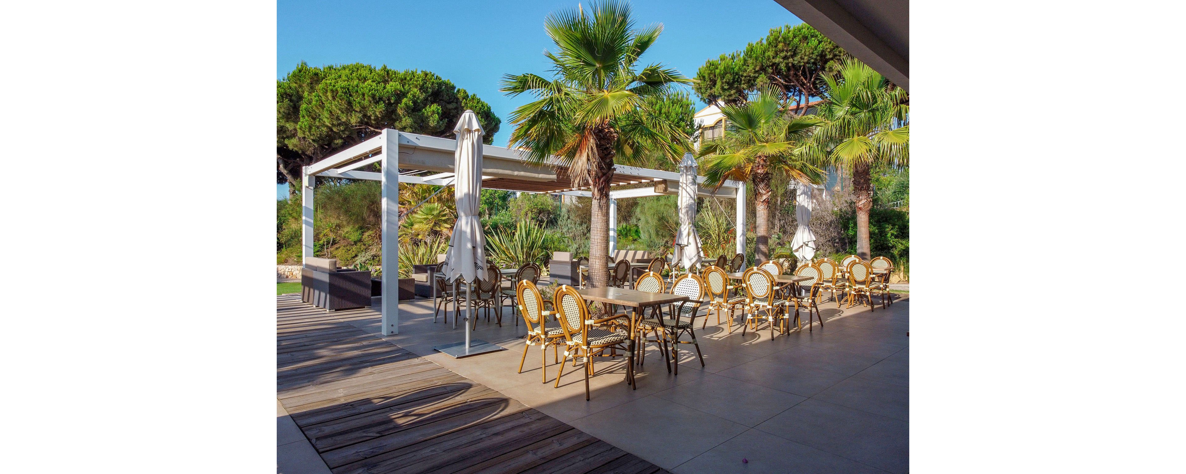 An open-air café terrace features neatly arranged tables and wicker chairs under a modern white pergola. Closed white umbrellas stand between the tables, suggesting early morning or off-hours. Tall palm trees and lush greenery surround the seating area, creating a relaxed, resort-like atmosphere. Warm sunlight and clear skies enhance the inviting, vacation-style setting.