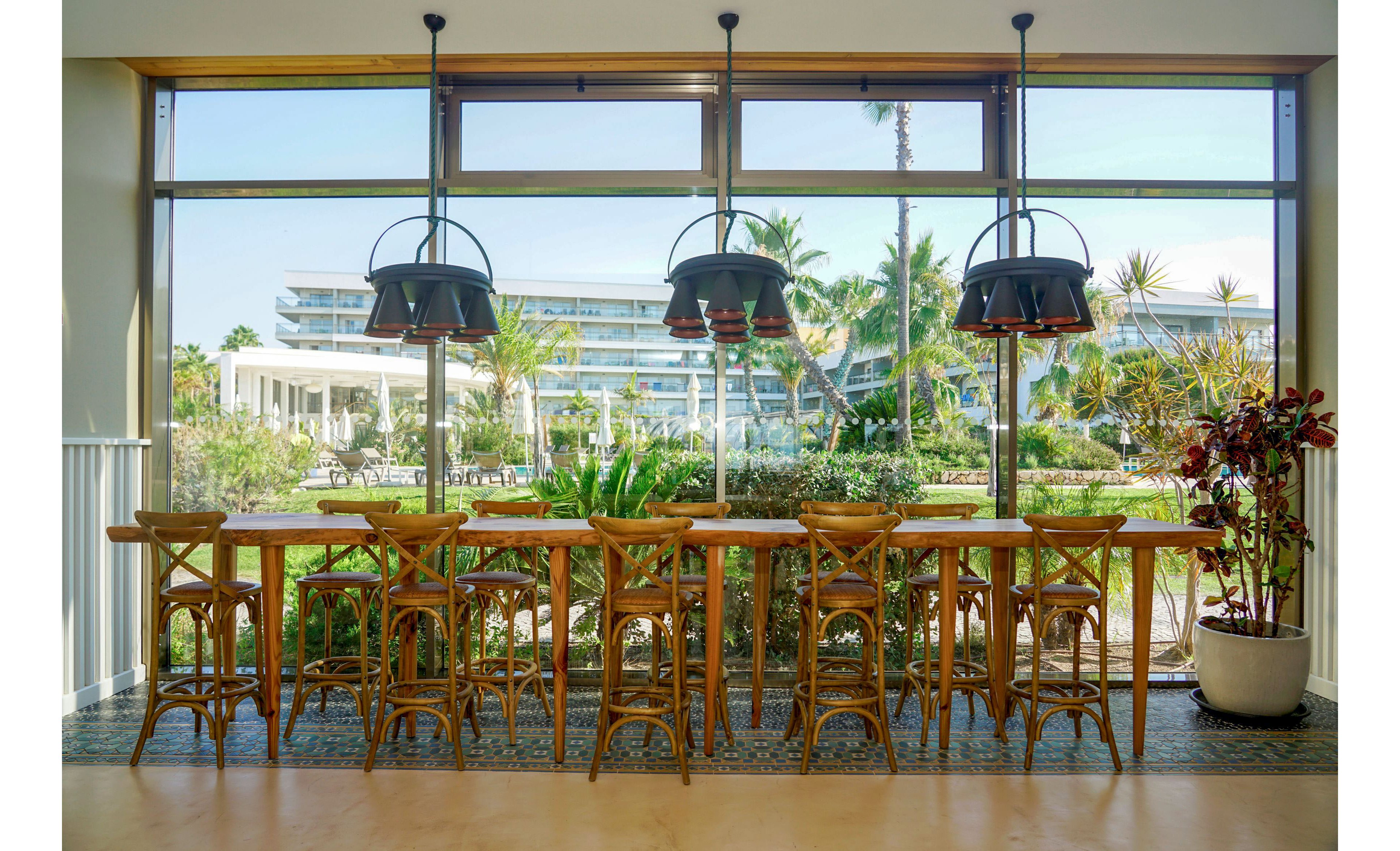 A long wooden bar-height table with matching stools is set in front of a large window in a bright café or restaurant. Black pendant lights hang above the table, while potted plants add greenery along the windowsill. Through the expansive glass, a modern resort complex, palm trees, and lush gardens are visible in the background. The scene feels airy and relaxed, suggesting a tropical vacation or coastal destination.