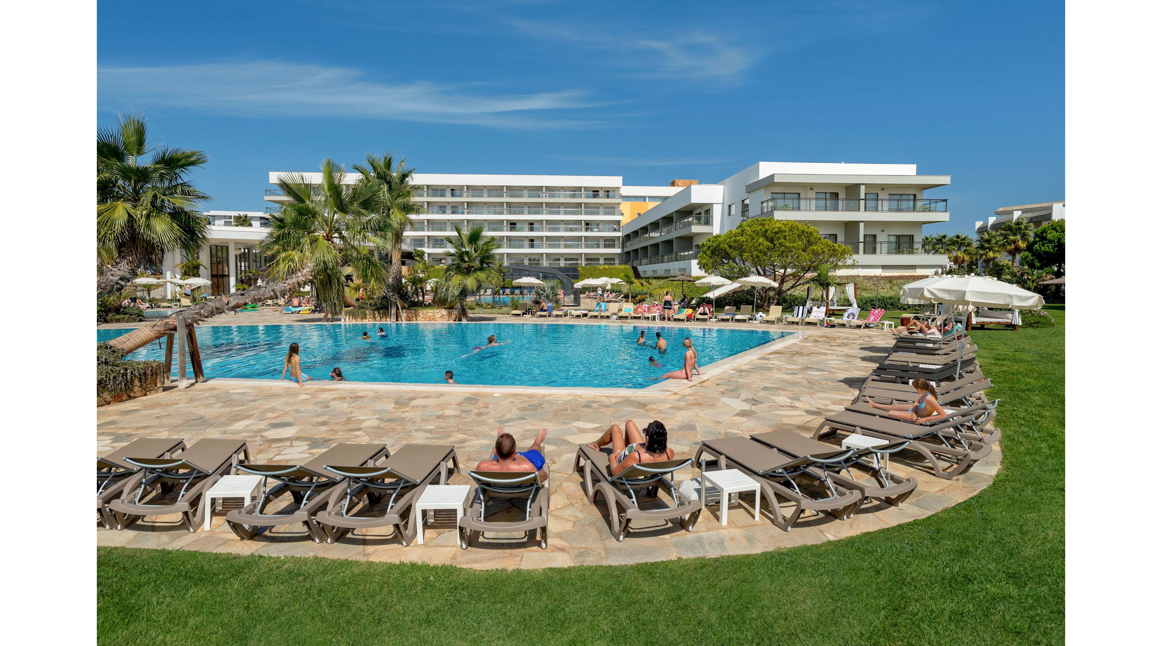 A large outdoor swimming pool is surrounded by numerous lounge chairs at a modern resort. Guests relax on sunbeds and swim in the clear blue water under a bright, cloudless sky. Palm trees and manicured lawns frame the pool area, adding a tropical feel. In the background, multi-story hotel buildings with balconies overlook the lively poolside scene.